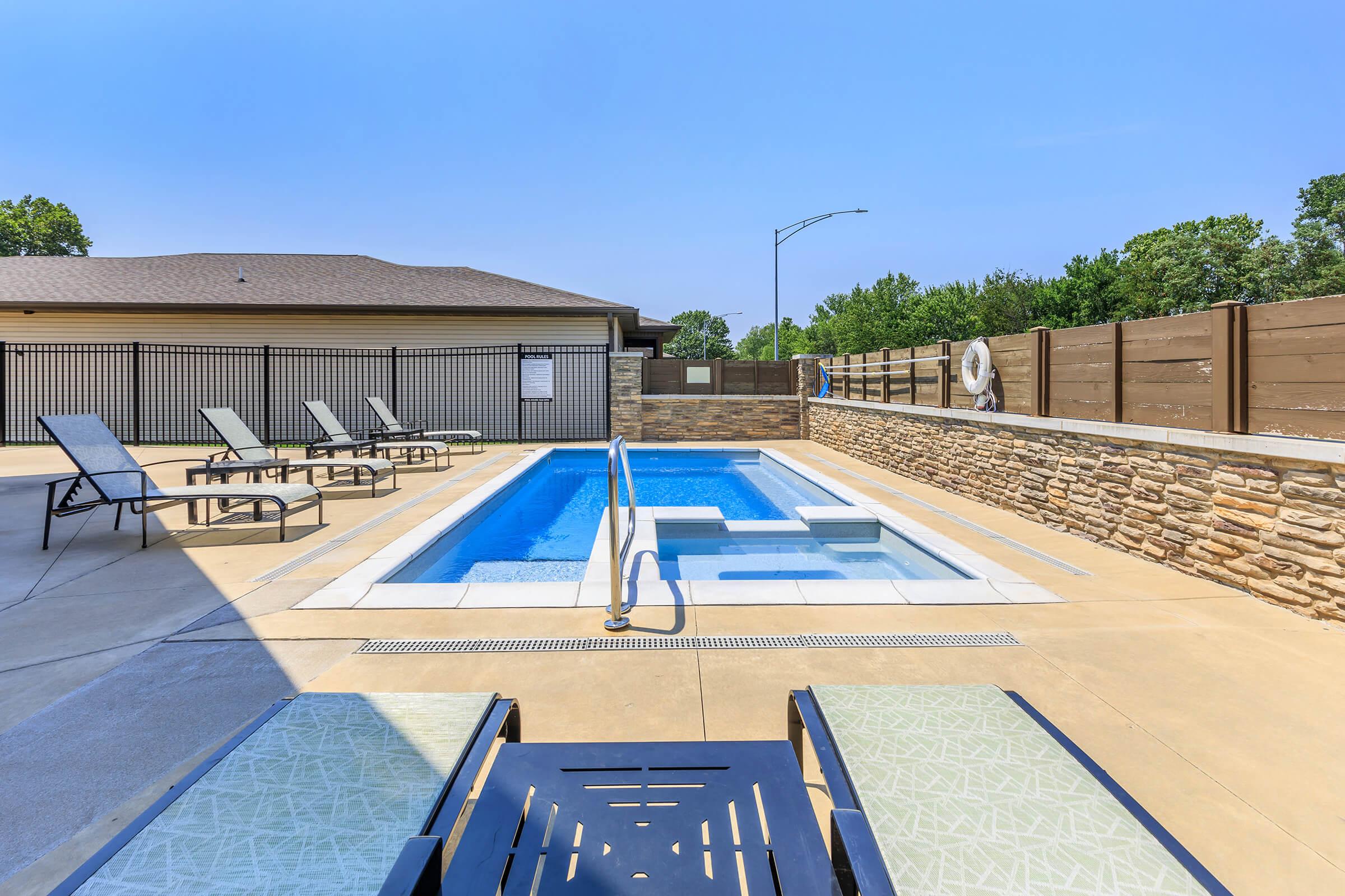 A serene outdoor pool area with a small swimming pool and a stone feature wall. Surrounding the pool are lounge chairs for relaxation, and a clear blue sky provides a bright backdrop. The area is enclosed with fencing, creating a private atmosphere.