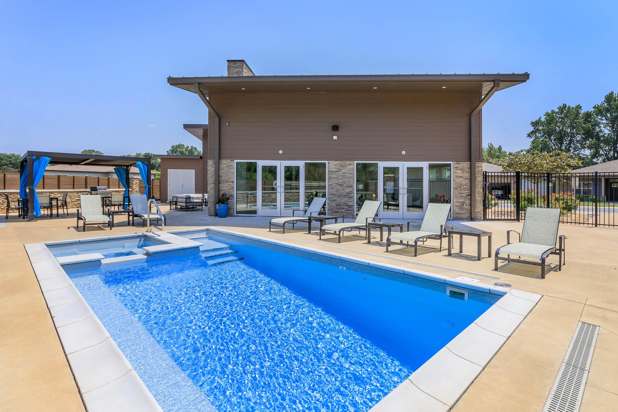 A modern outdoor pool area featuring a sparkling blue pool with steps, surrounded by lounge chairs. In the background, there's a contemporary building with large windows, flanked by landscaping and a clear blue sky. A shaded seating area is visible to the left.