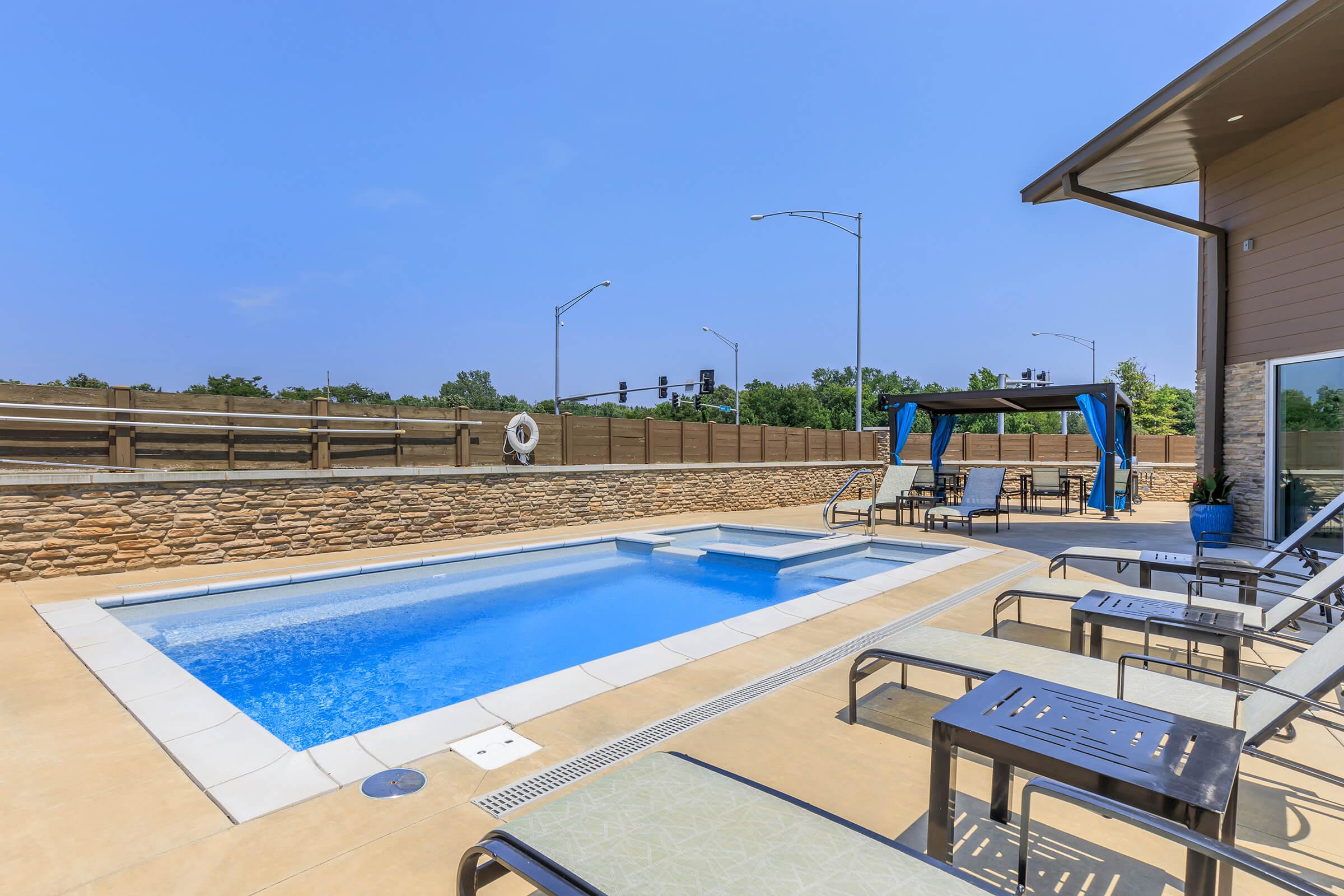 A sunny outdoor area featuring a small pool surrounded by lounge chairs. There is a shaded seating area with a canopy and a backdrop of a wooden fence. The scene includes nearby streetlights and a clear blue sky, suggesting a relaxing atmosphere for leisure activities.