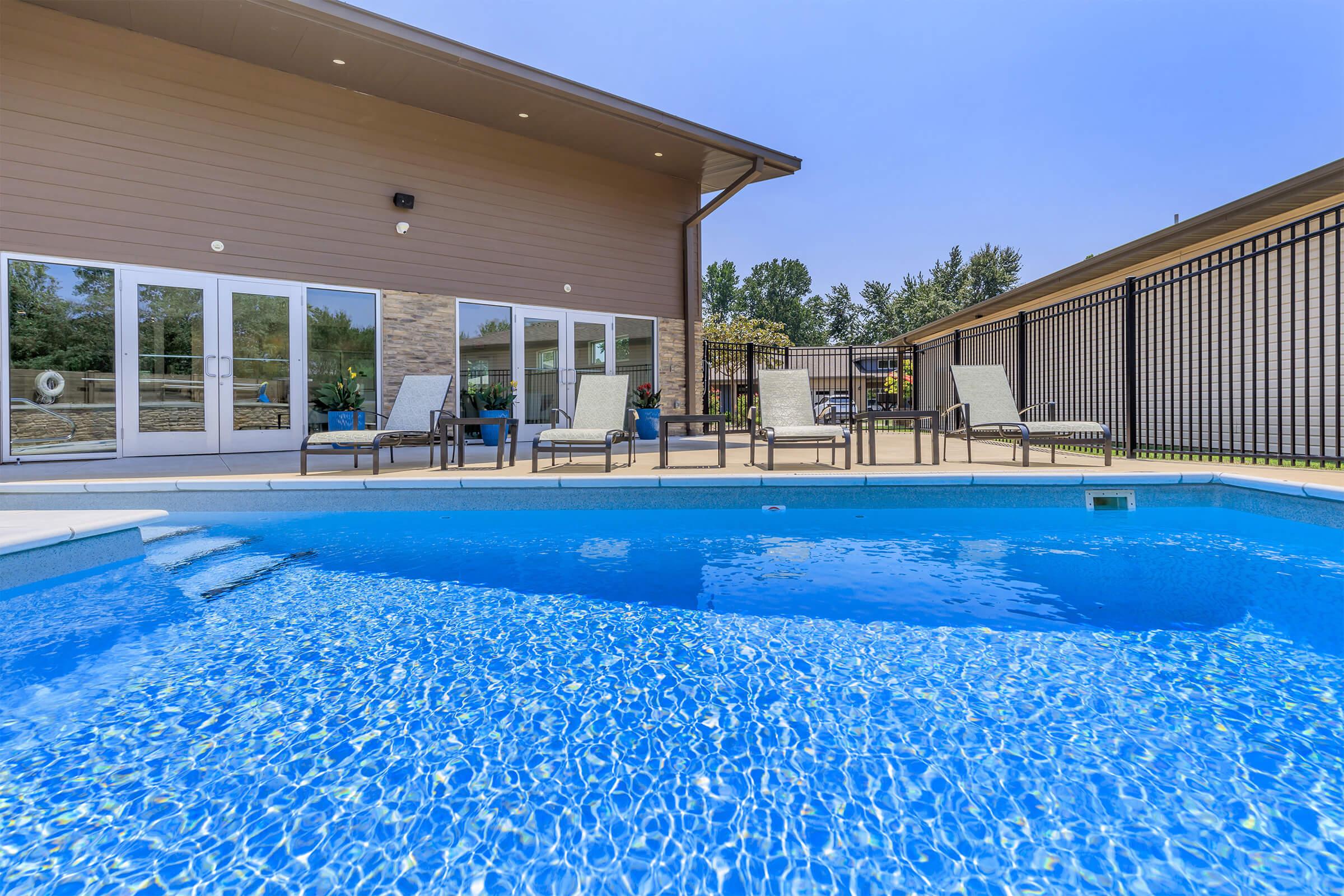 A modern outdoor area featuring a swimming pool with clear blue water. In the background, there is a building with large windows and a covered patio. Lounge chairs line the poolside, and the surrounding landscape includes trees and green grass. The sky appears clear and bright.