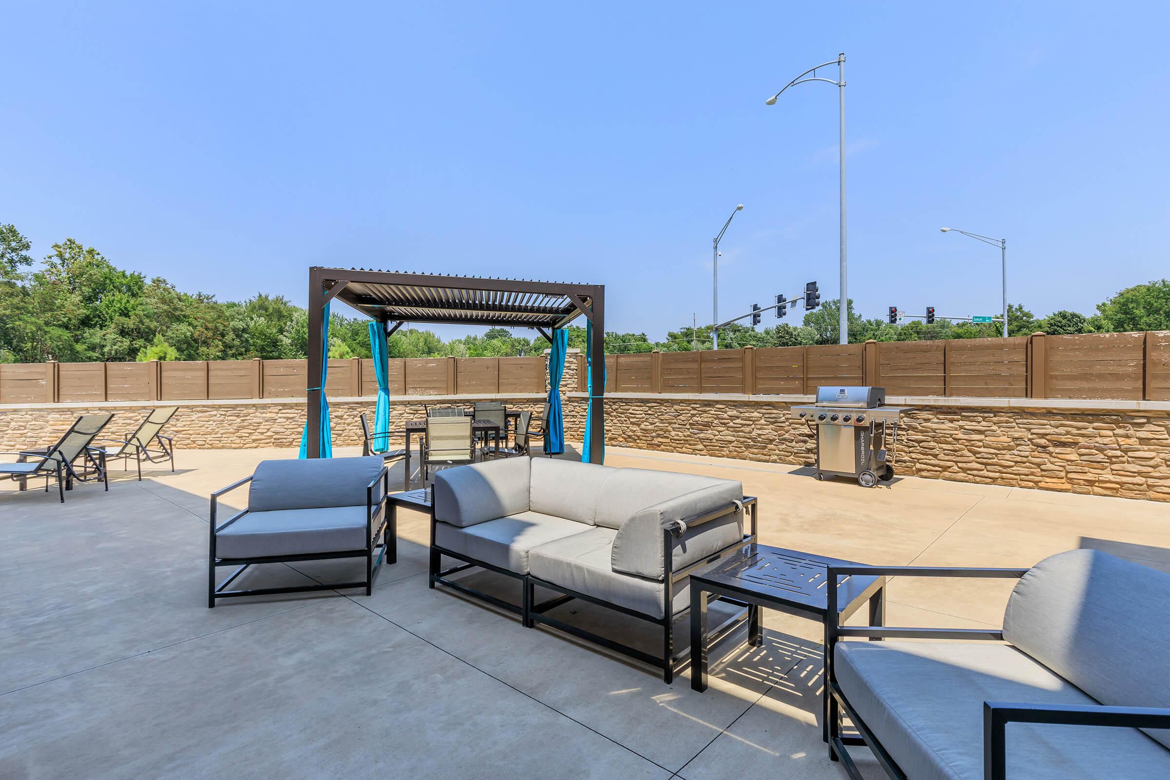 An outdoor lounge area featuring modern seating, a shaded pergola, and a grill. The space is surrounded by a stone wall and has various trees in the background under a clear blue sky. Traffic lights and a road are visible in the distance.