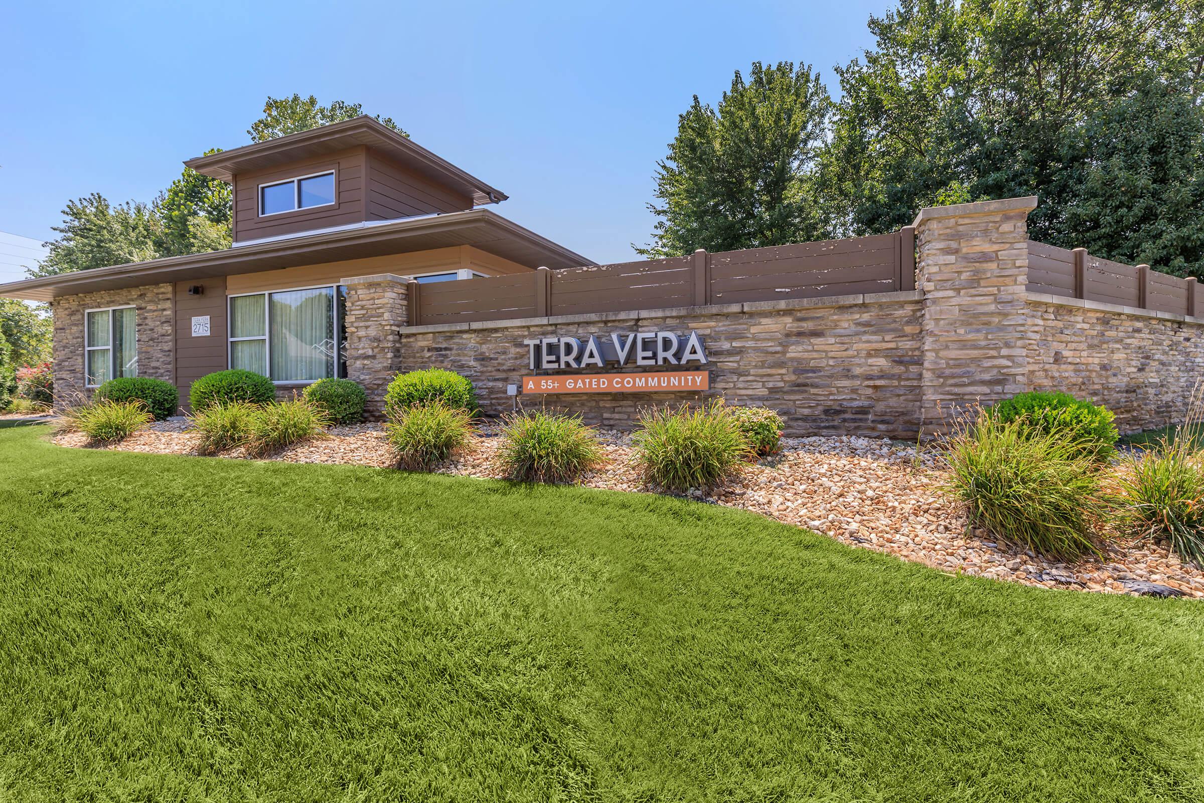 Sign for "Tera Vera," a 55+ gated community, displayed on a stone wall, surrounded by landscaped greenery, shrubs, and rocks. The building behind is modern with large windows, set against a clear blue sky and trees in the background.
