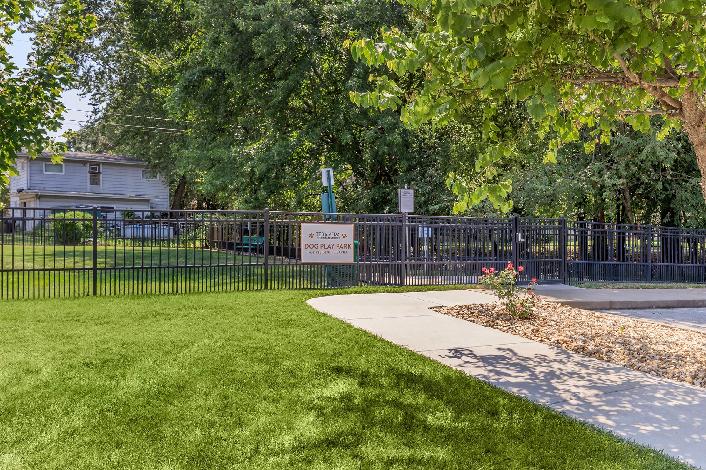 A fenced dog play park featuring green grass, a pathway, and signage indicating the park's name. Nearby trees provide shade, and there are benches visible in the background. The area is well-maintained, offering a welcoming environment for dogs and their owners.