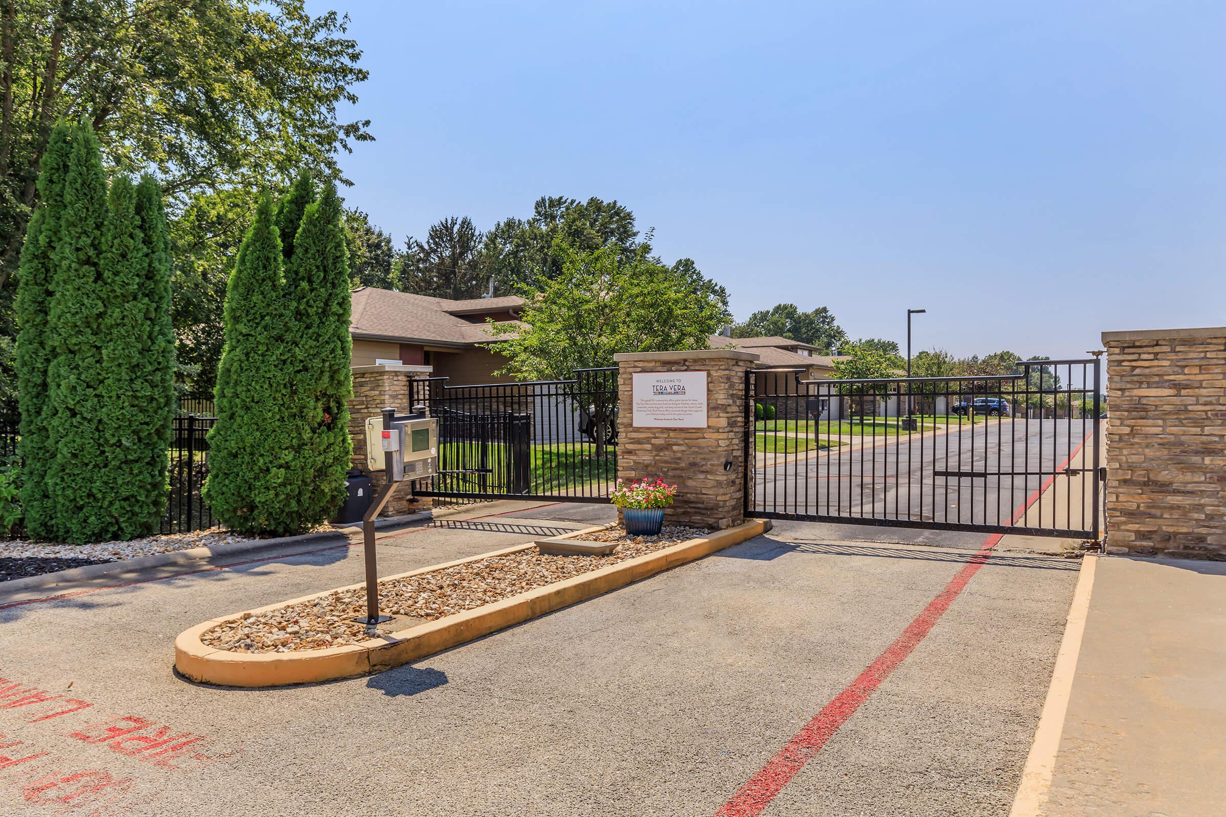 A gated entrance with a decorative mailbox and a flower pot, flanked by tall green shrubbery and trees. The drive is paved and leads into a residential area, with a clear blue sky visible above. A sign on the gate indicates the property's name or access information.