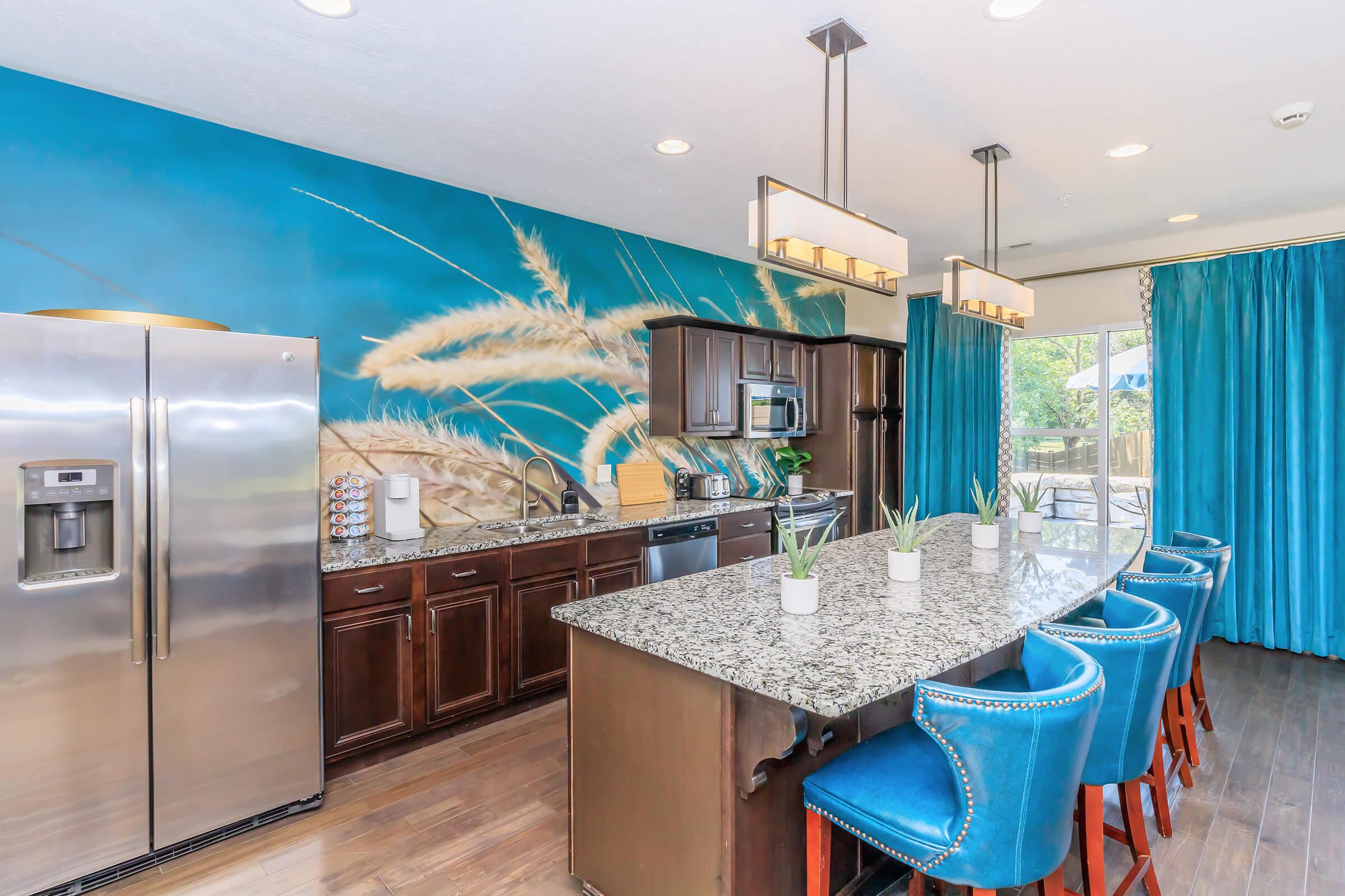 Modern kitchen featuring a granite island with blue upholstered bar stools, stainless steel appliances, and wooden cabinetry. A large mural of wheat on the wall adds a natural touch, while bright turquoise curtains frame the window, enhancing the light and airy atmosphere.