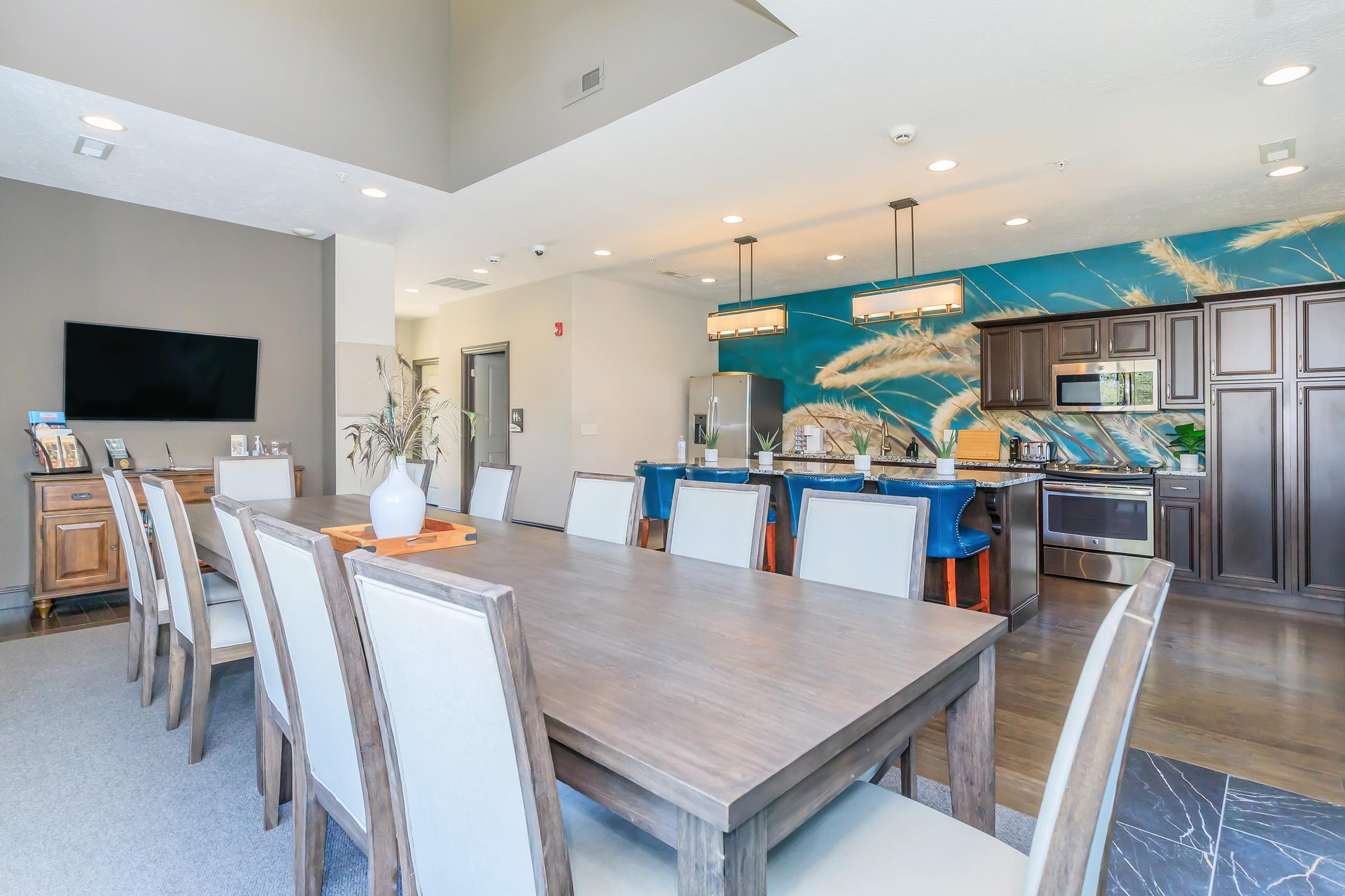 Modern dining area featuring a long wooden table with beige chairs, set against a backdrop of a stylish kitchen. The kitchen includes dark wood cabinets, stainless steel appliances, and a vibrant blue wall with decorative elements. A large flat-screen TV is mounted on the wall, adding to the contemporary ambiance.