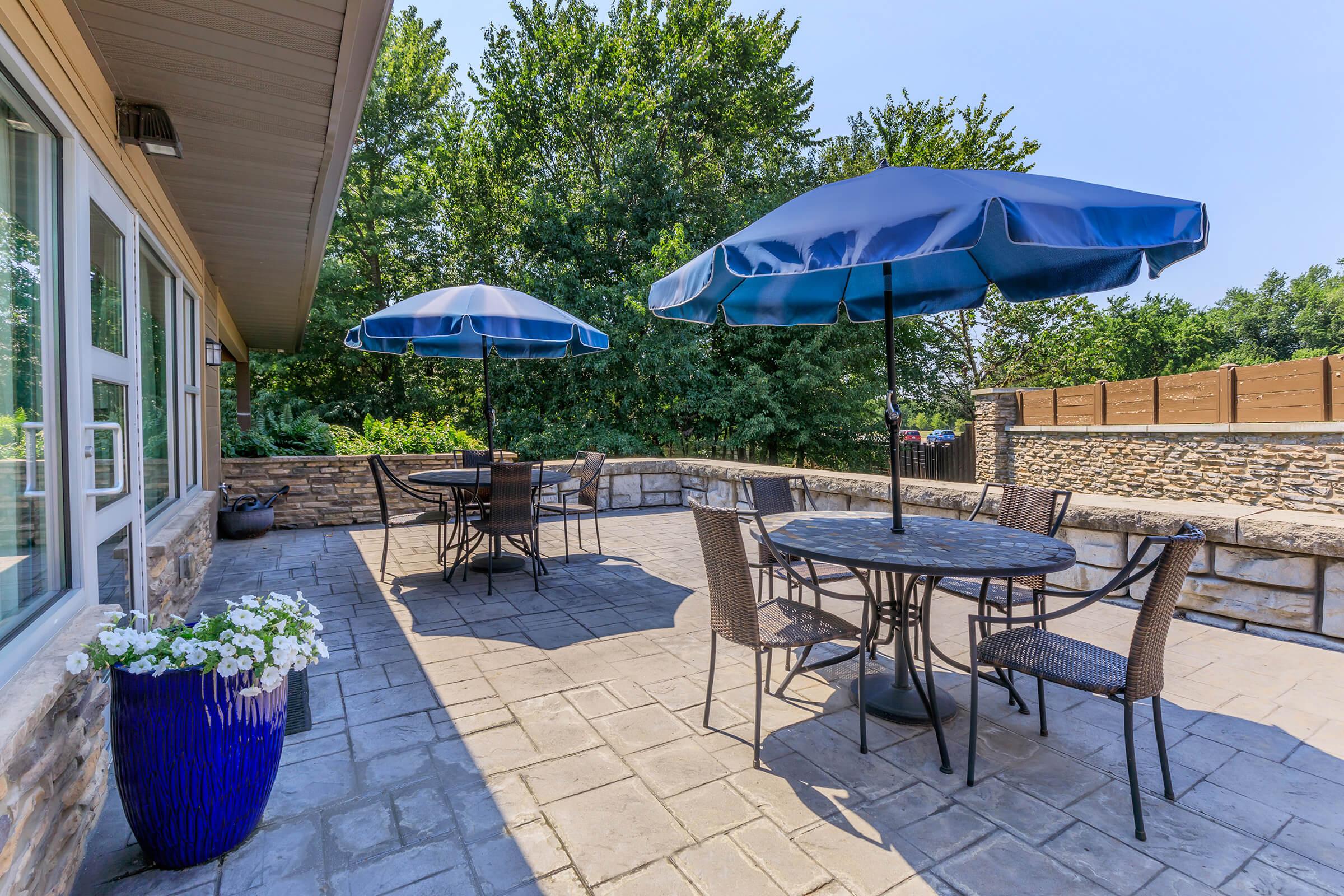 A sunny outdoor patio featuring two blue umbrellas over round tables with chairs. The stone wall and paved floor complement the lush greenery in the background. A blue planter holds white flowers, adding a touch of color to the inviting space. Perfect for relaxation or outdoor dining.
