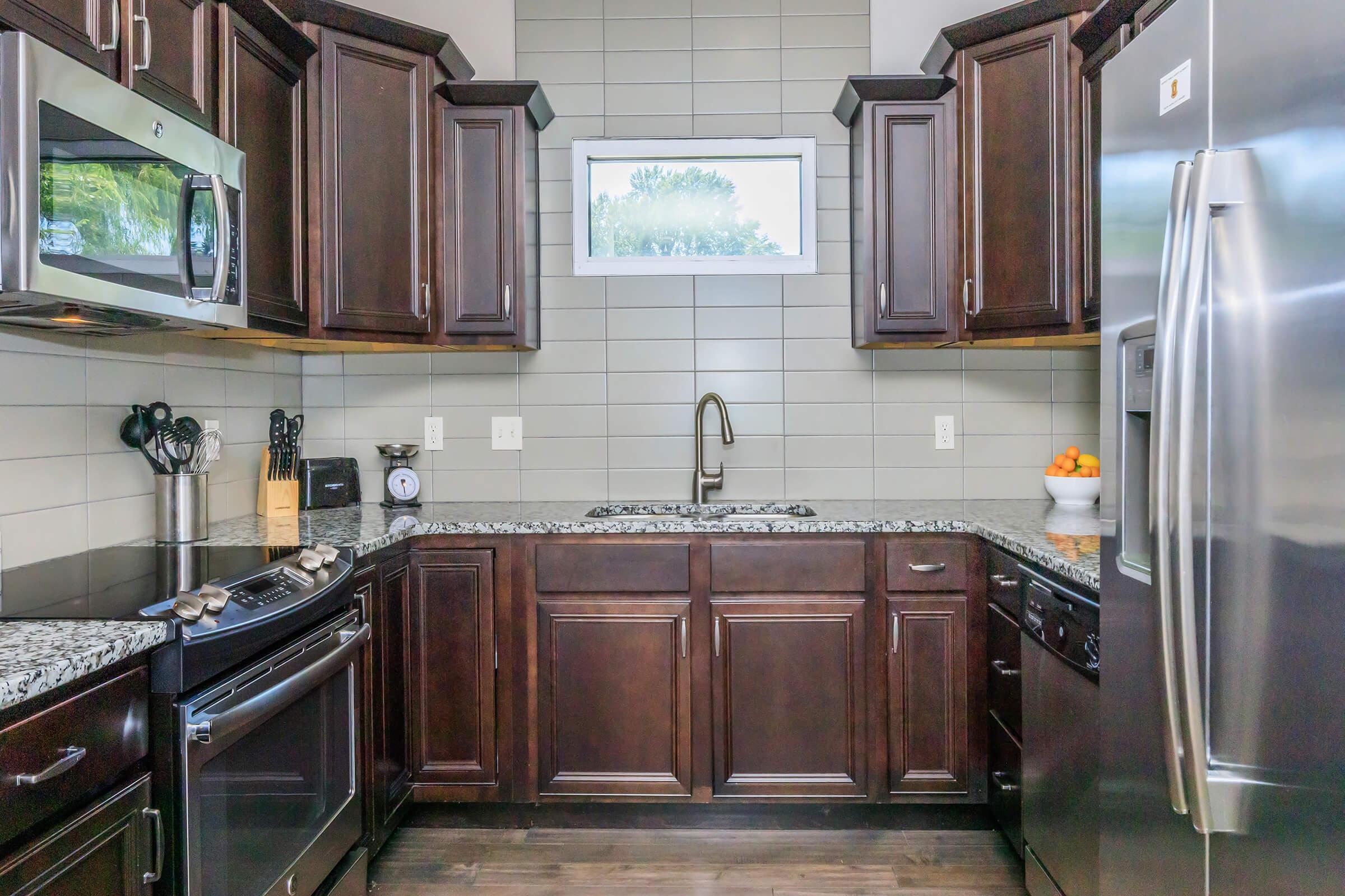 Modern kitchen featuring dark wood cabinets, a stainless steel refrigerator, a microwave, and a gas stove. The backsplash has gray tiles, and the countertop is made of granite. A window above the sink lets in natural light, and a bowl of oranges sits on the countertop.