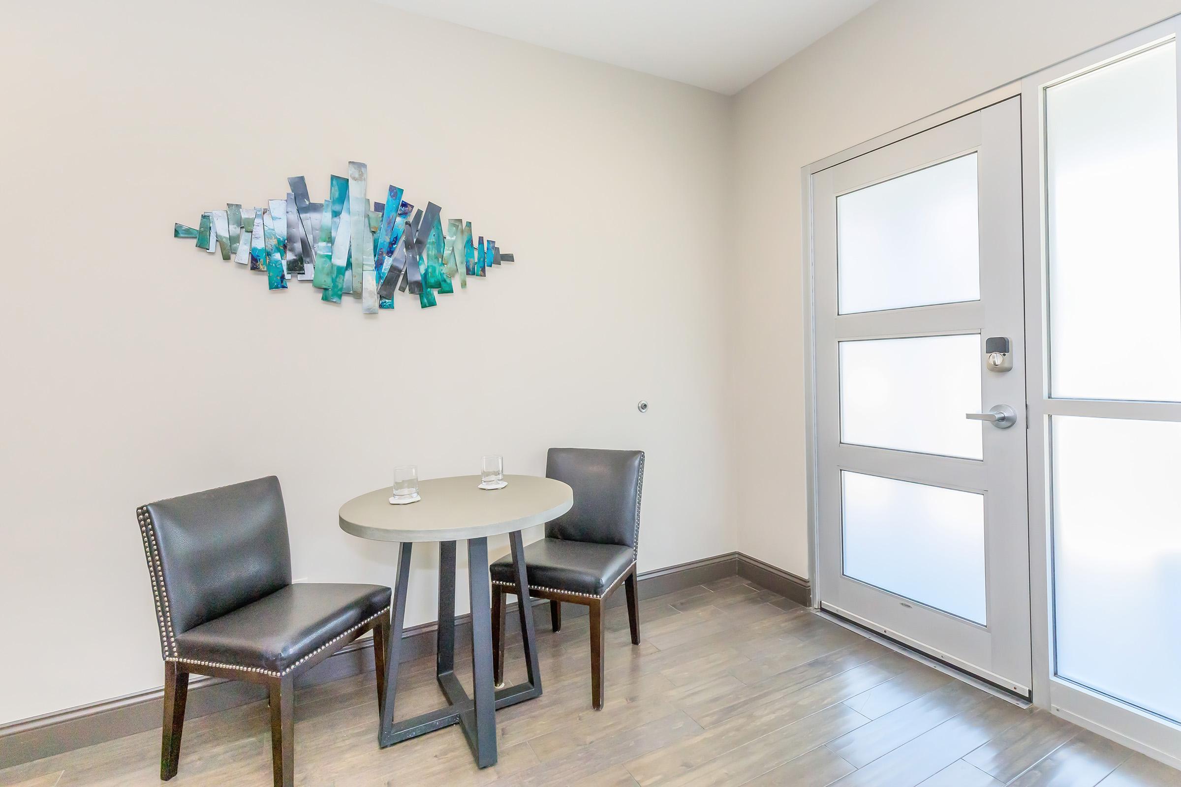 A minimalist room featuring a small round table with two chairs. Glasses are placed on the table. A decorative wall piece in shades of blue and green is mounted on a light-colored wall. There's a modern door with multiple panels leading outside, complementing the contemporary design.