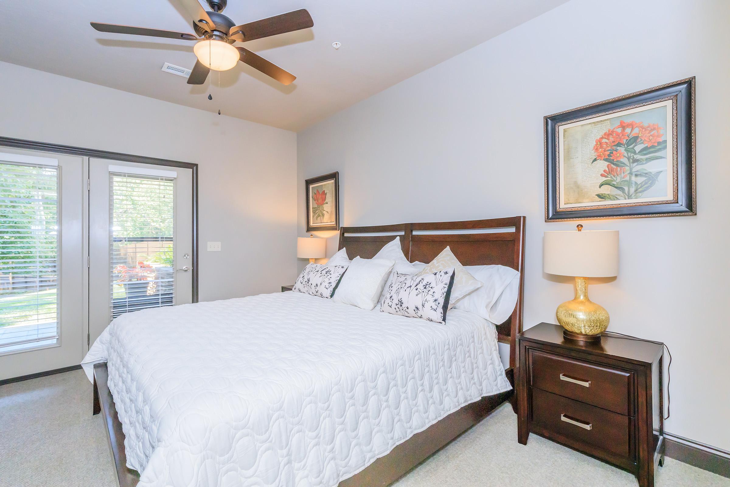 Cozy bedroom featuring a queen-sized bed with a white quilt and decorative pillows. There's a wooden bedside table with a gold lamp on one side and framed floral artwork on the walls. A ceiling fan adds to the ambiance, and large windows provide natural light and a view of the outdoors.