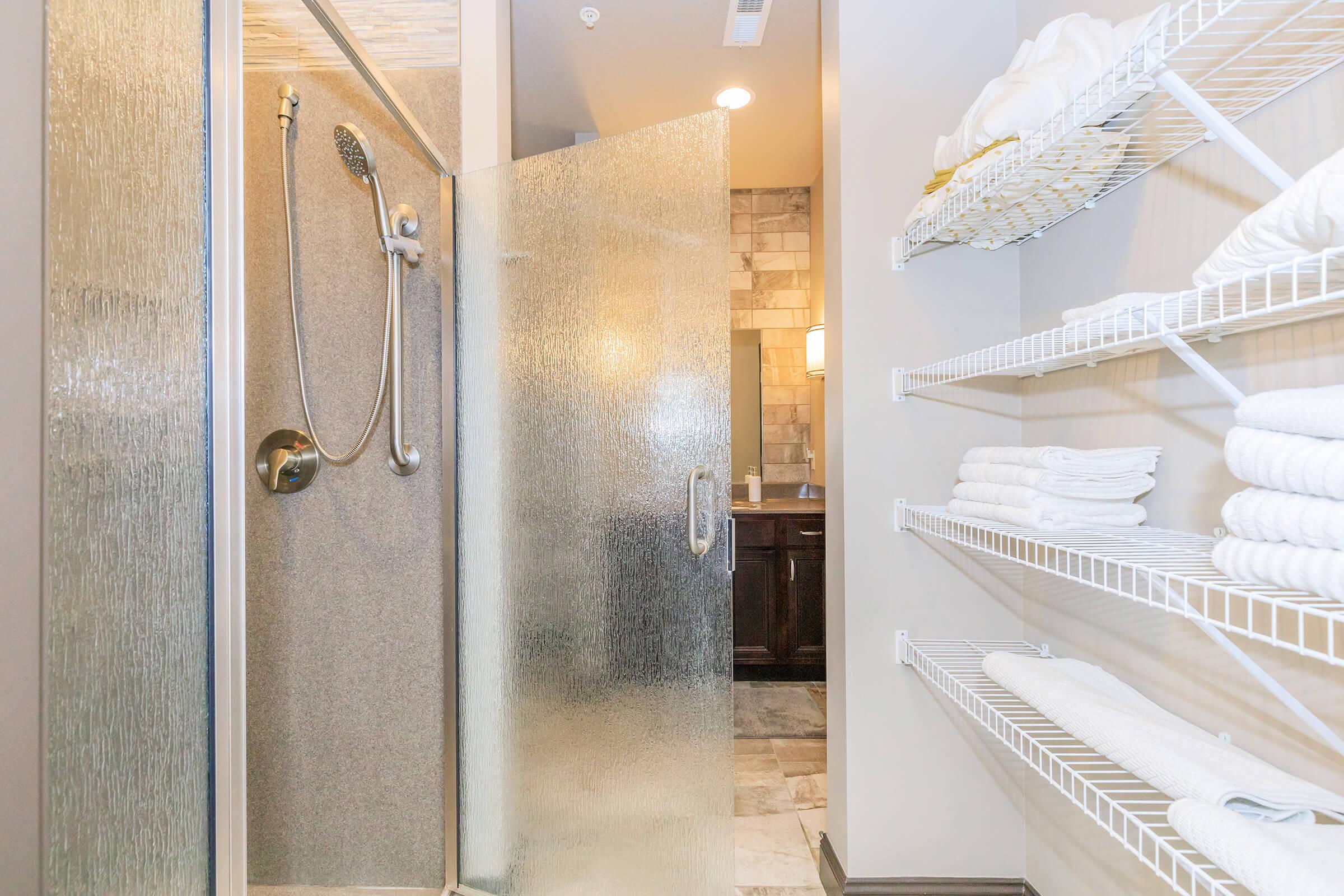A modern bathroom featuring a glass shower with a handheld showerhead, alongside neatly organized shelves stocked with white towels. The walls are finished in neutral tones with stone accents, and a vanity can be seen in the background, softly lit for a warm ambiance.