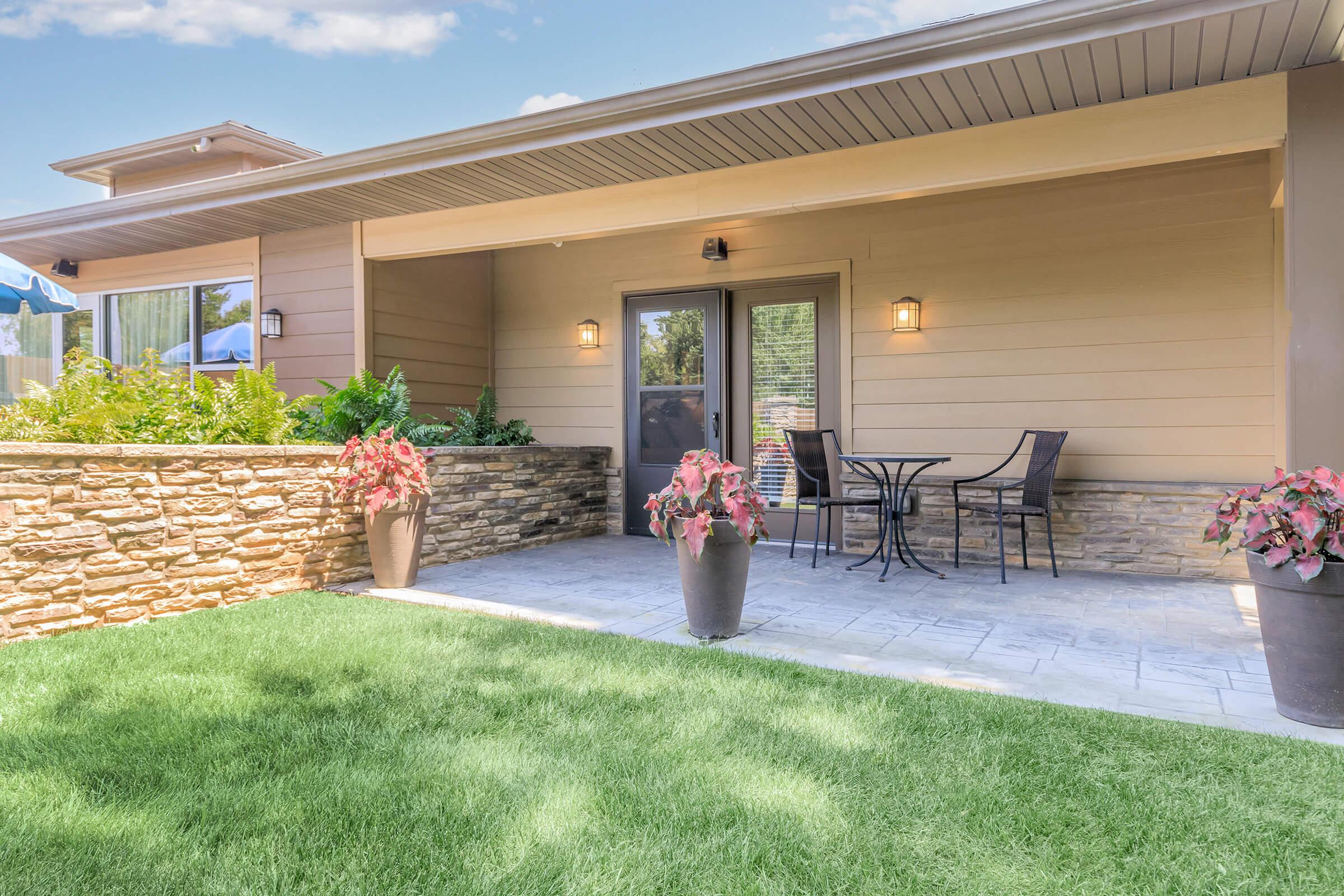 A modern patio area featuring a stone wall, potted flowers, and a small table with two chairs. The patio is surrounded by green grass and offers a bright, inviting atmosphere with warm lighting and a clear blue sky in the background.