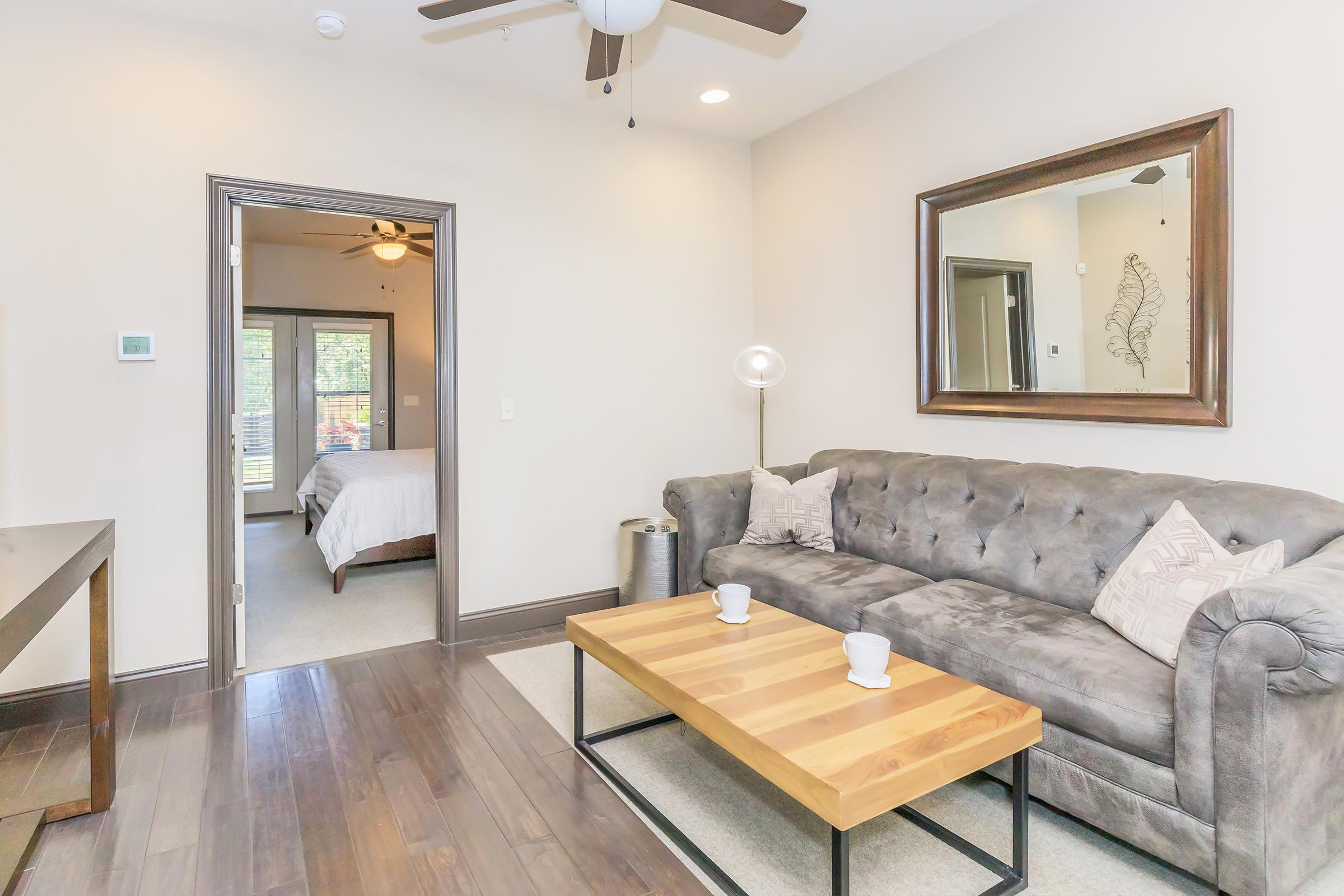 A stylish living room featuring a gray tufted sofa with two cups on a wooden coffee table. A decorative mirror hangs on the wall. In the background, a doorway leads to a bedroom with two ceiling fans visible. The room has warm, neutral tones and hardwood flooring, creating a cozy atmosphere.