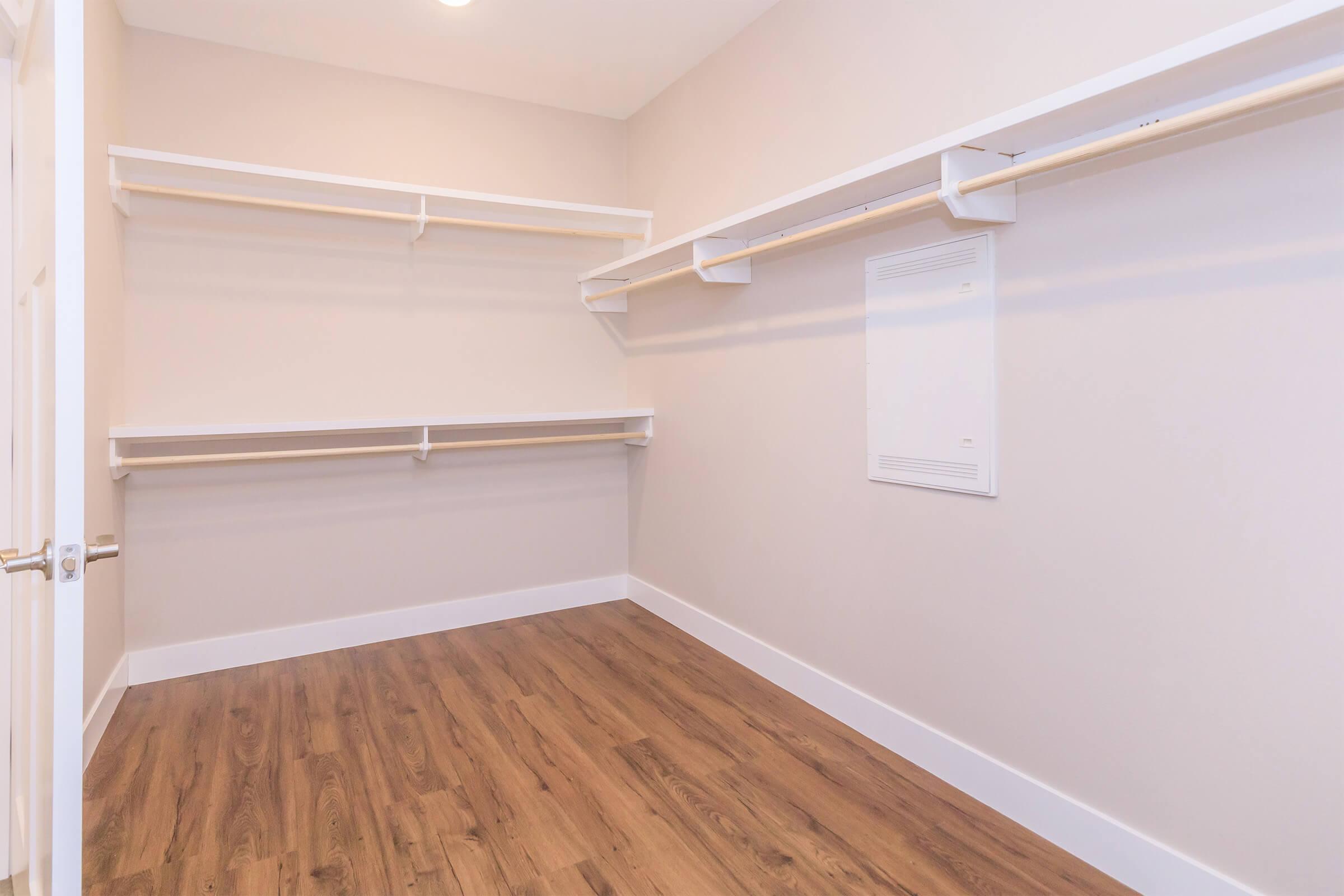 A clean, empty closet featuring two hanging rods mounted on white walls. The floor is finished with light wood laminate, and there is a small electrical panel on one wall. The overall space is bright and uncluttered.