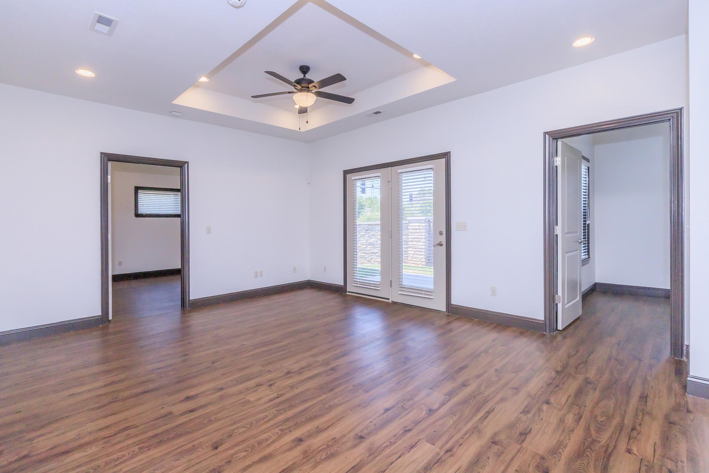 Spacious interior of a modern living room featuring wood flooring, a ceiling fan, and large windows. Double doors lead to an outdoor area, while entrances to other rooms are visible on the left and right. The walls are painted in a light color, enhancing the bright and airy atmosphere.