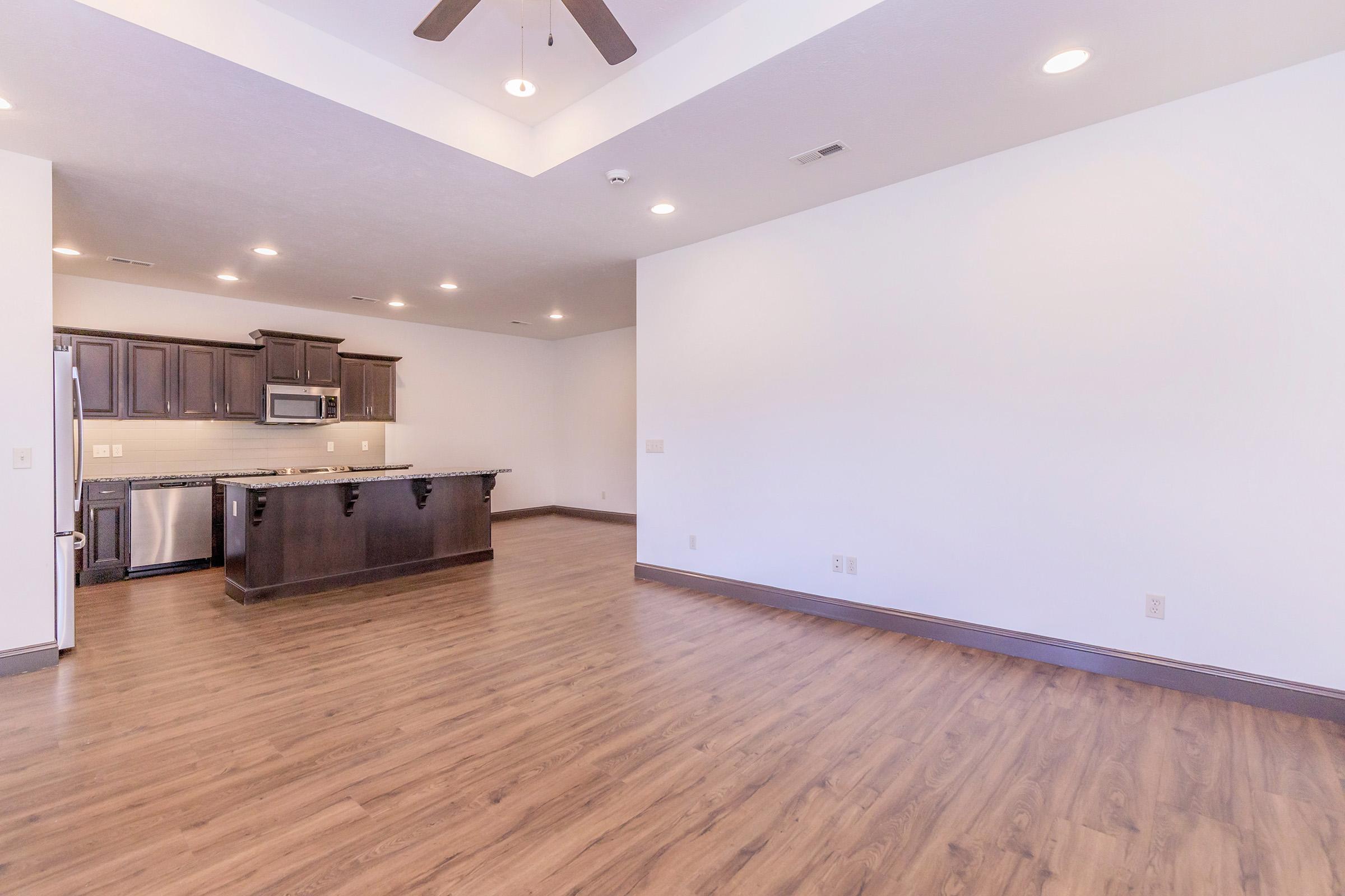 Spacious interior of a modern kitchen and living area, featuring dark wood cabinets, stainless steel appliances, and an island. The living space has wood-like flooring and a ceiling fan, with neutral-colored walls and bright lighting, creating an inviting atmosphere.