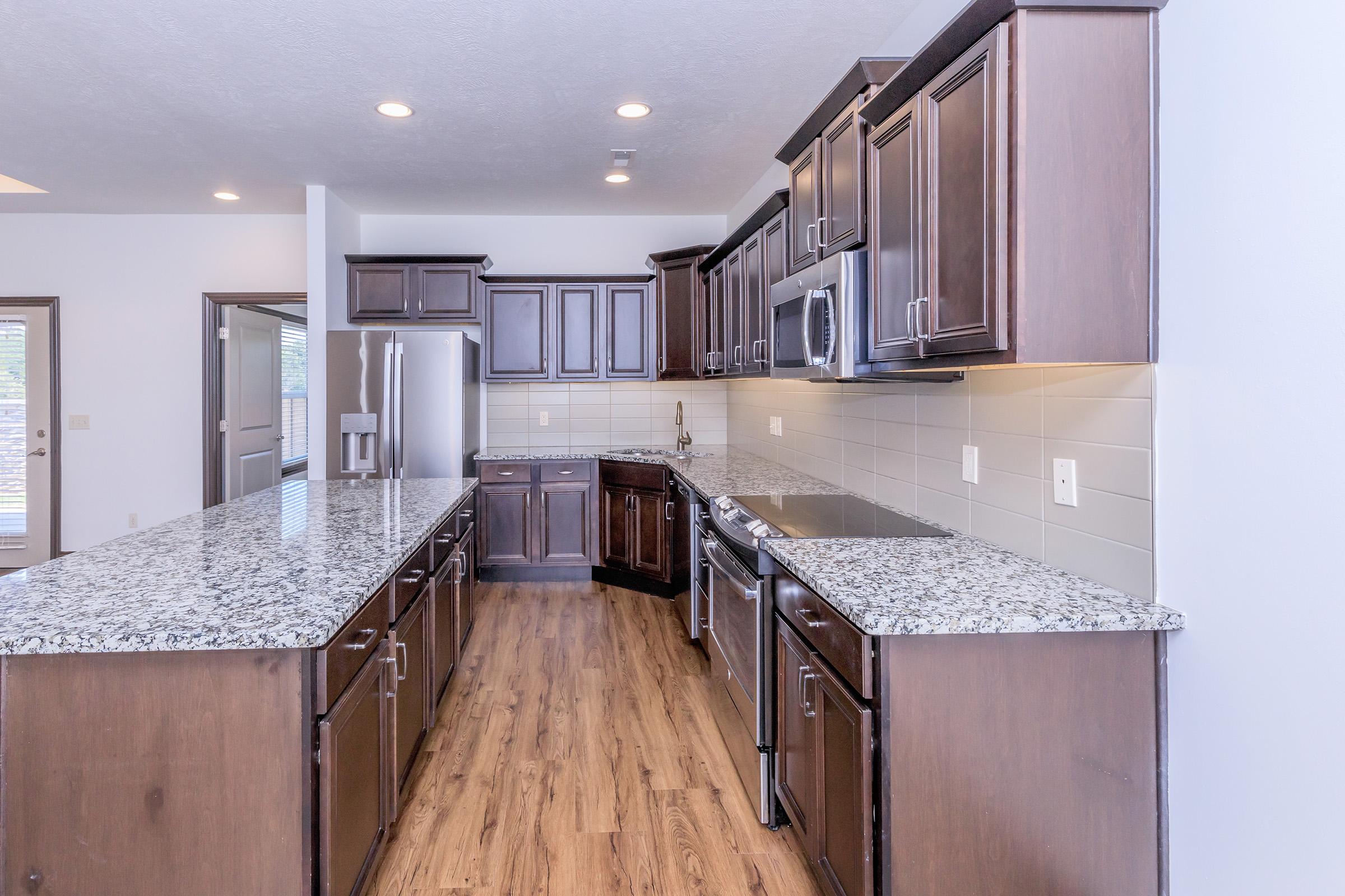 Modern kitchen featuring dark wood cabinets, stainless steel appliances, and a large granite countertop. The space is well-lit with recessed lighting and has a warm wood laminate floor, creating an inviting atmosphere. Large windows allow natural light to fill the room.