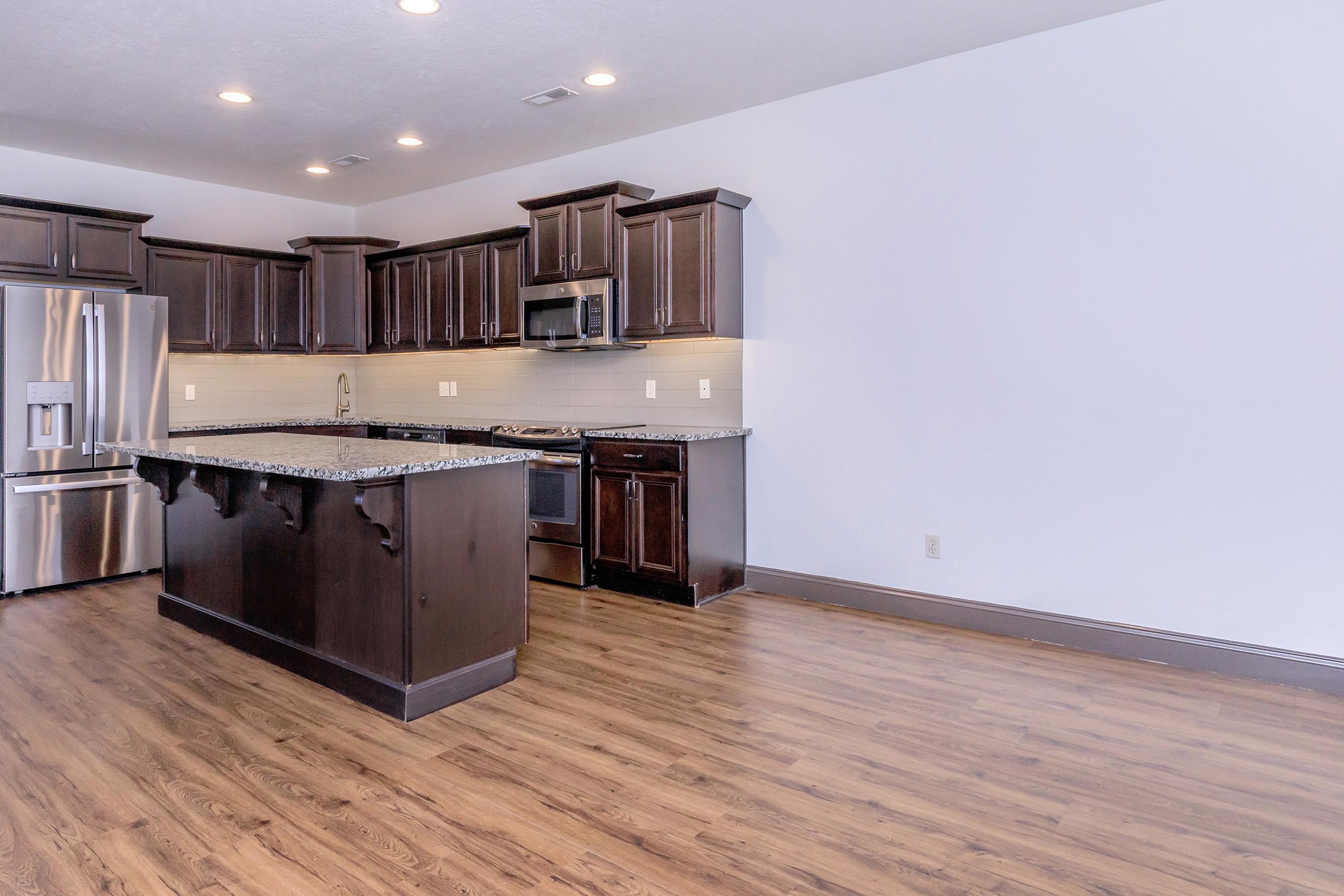 Modern kitchen featuring dark wooden cabinets, stainless steel appliances including a refrigerator and oven, a granite countertop island, and light-colored tile backsplash. The floor is covered in wood-like laminate, and the walls are painted white, creating a clean and airy atmosphere.