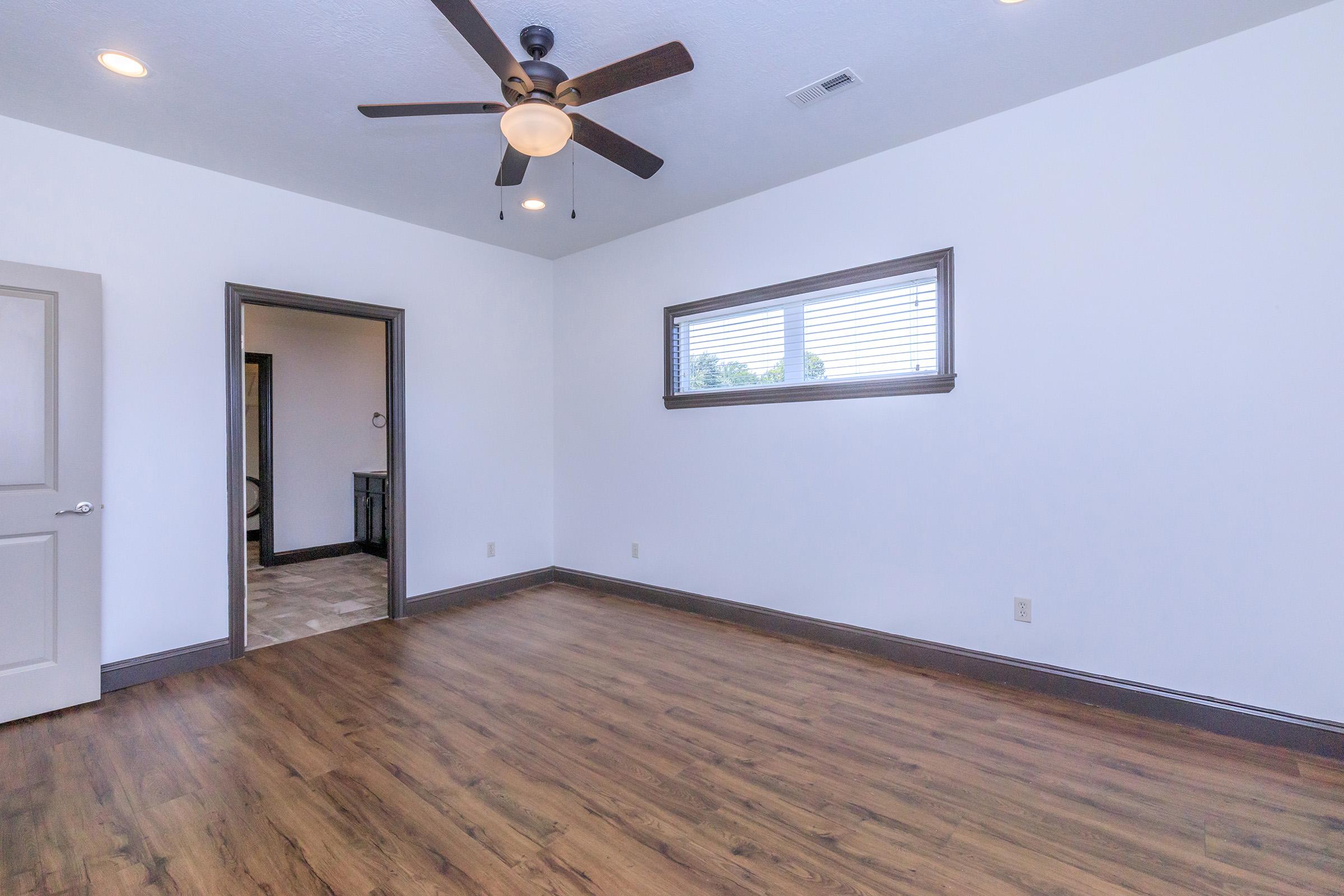 A vacant room featuring a wooden floor, a ceiling fan with five blades, and a window with blinds. There is a doorway leading to another room on the left, and the walls are painted white with dark gray trim. The space is well-lit, showcasing a minimalist design without furniture.