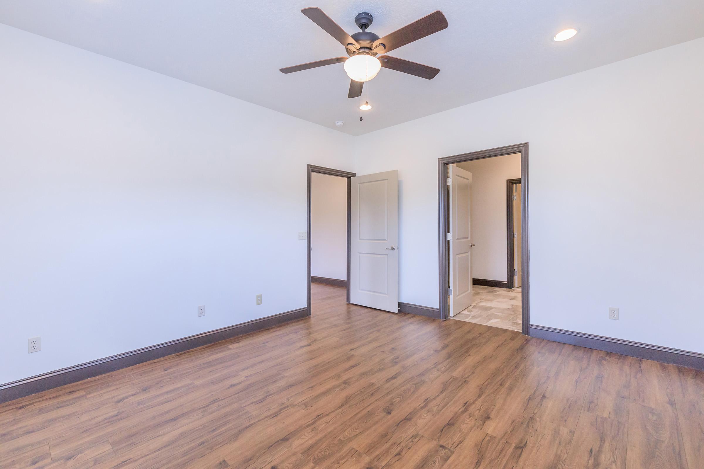 A spacious, empty room with light-colored walls and a ceiling fan. The floor features wood-like laminate, and there are doorways leading to other rooms visible. The lighting is bright, creating an open and airy atmosphere.