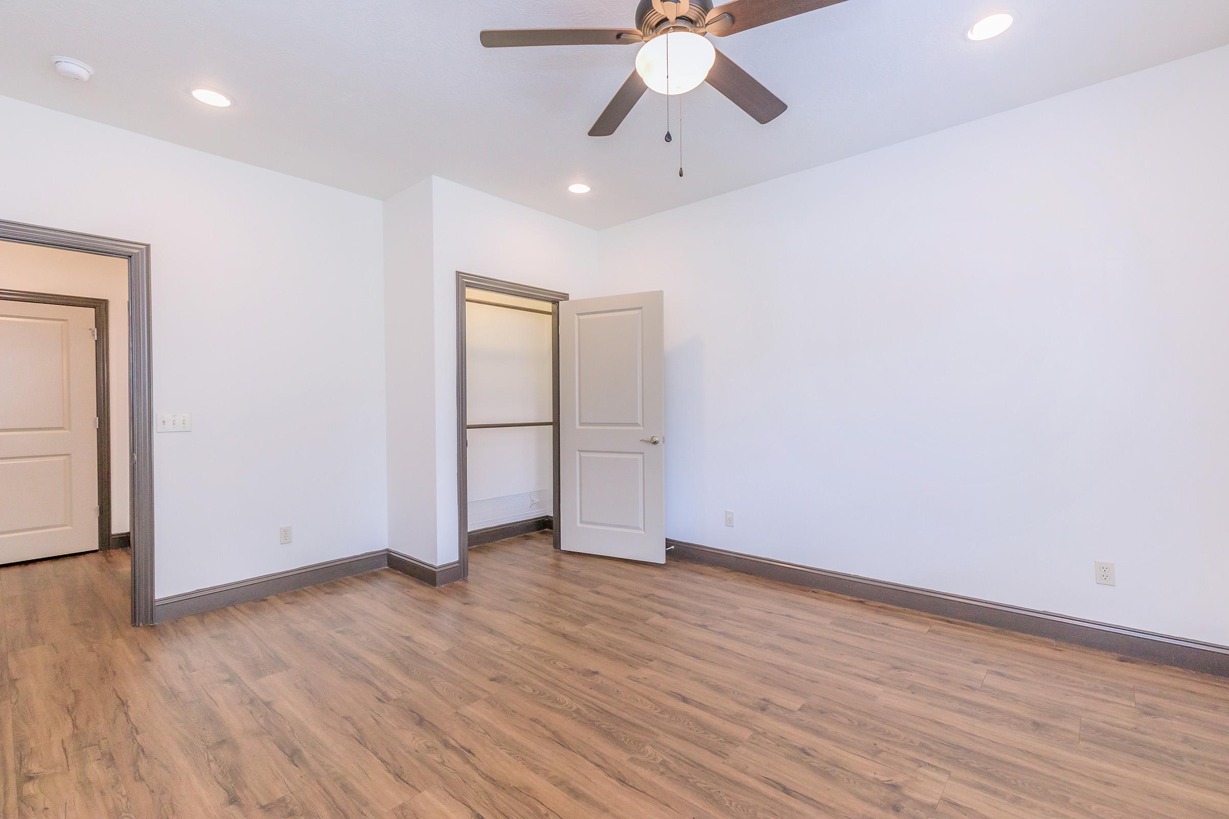 A spacious, empty room featuring light hardwood flooring, a ceiling fan, and white walls. There are two doorways: one leading to a closet with an open door and another leading to a closed door. The room is well-lit with natural light. Ideal for various design possibilities.