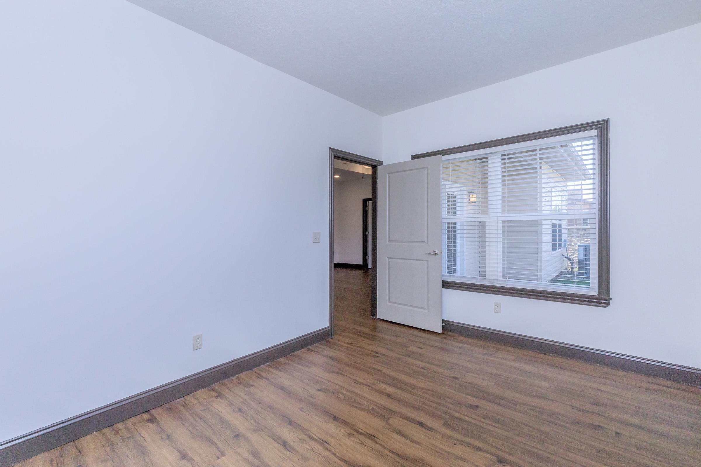 An empty room with white walls, hardwood flooring, and a partially open door leading to another space. Natural light streams in through a window featuring horizontal blinds. The room is unadorned, providing a neutral backdrop for various uses.