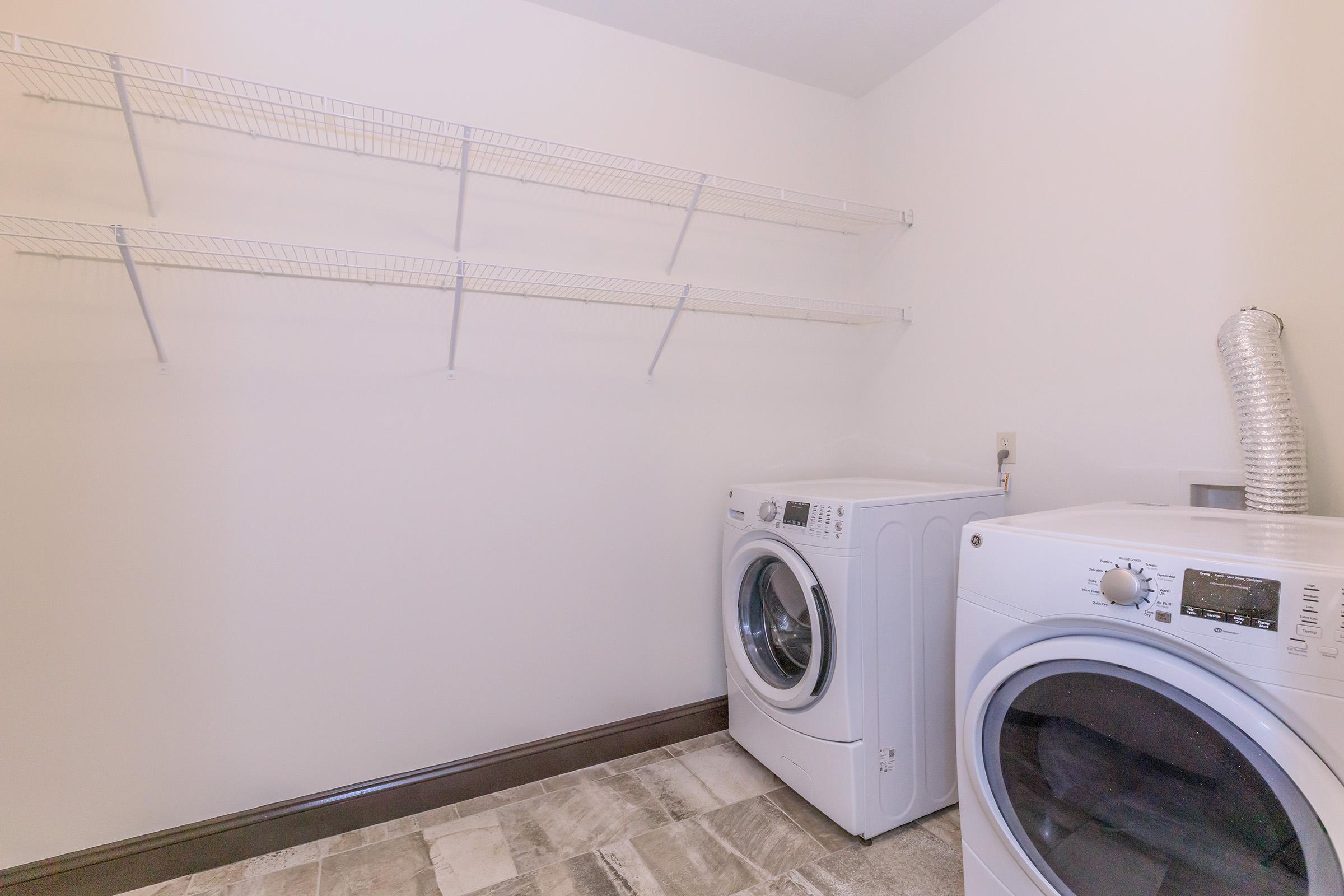A clean laundry room featuring two white washing machines side by side, with a wire shelving unit mounted on the wall above. The floor is tiled in a neutral color, and there is a dryer vent visible on the wall. The overall space is bright and uncluttered.