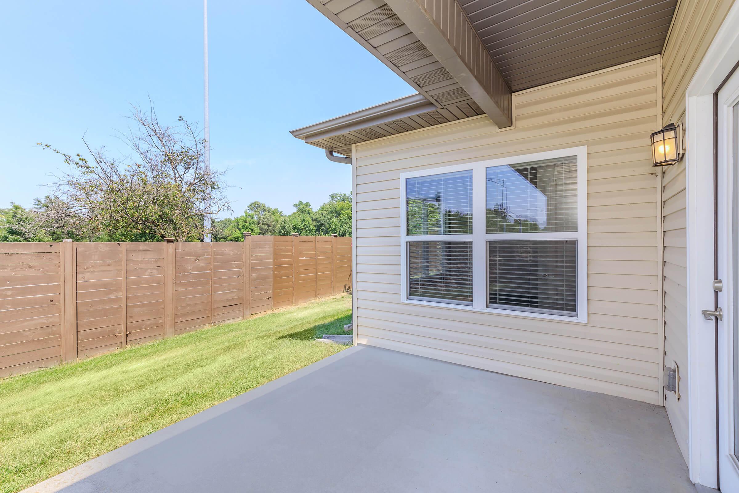 A view of a residential patio area featuring a beige exterior wall, large windows with white trim, and a well-maintained grassy yard. A wooden fence outlines the space, and a clear blue sky is visible above. The setting appears bright and inviting, ideal for outdoor relaxation.