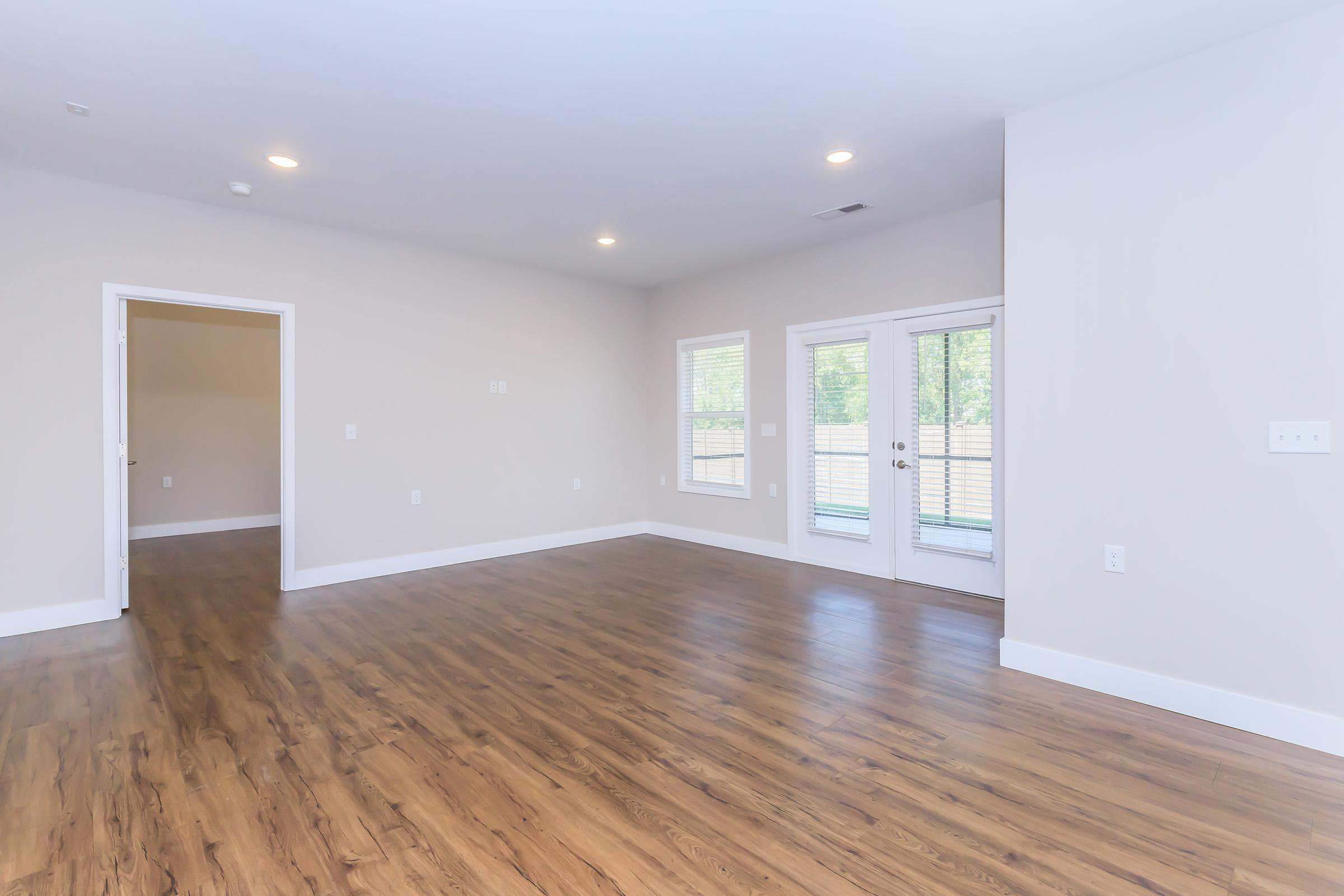 Bright interior of a spacious room with light-colored walls and wooden flooring. There are two doors: one leading to another room and the other to a small outdoor area. Large windows allow natural light to fill the space, creating a welcoming atmosphere.