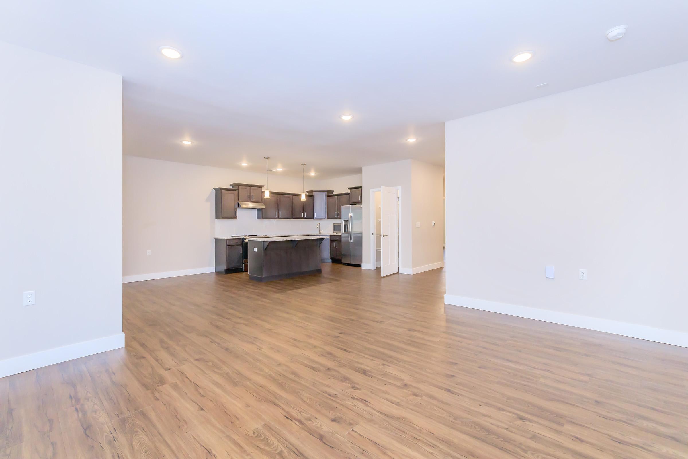 Spacious interior of a modern kitchen and living area featuring hardwood floors, abundant natural light, and minimalist décor. The kitchen includes dark cabinetry and stainless steel appliances, with an open layout connecting to the living space. Neutral wall colors enhance the airy, inviting atmosphere.