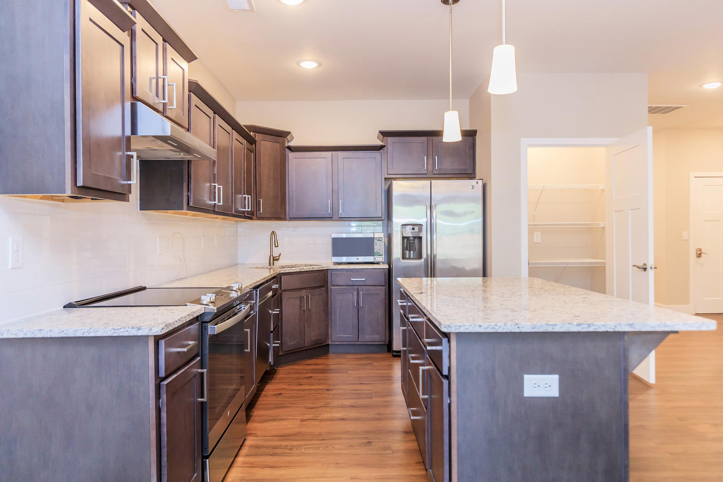 Modern kitchen featuring dark wood cabinetry, granite countertops, stainless steel appliances, and an island with seating area. There are pendant lights above the island, and a pantry door is visible in the background. The flooring is a warm wood tone, contributing to a stylish and functional space.
