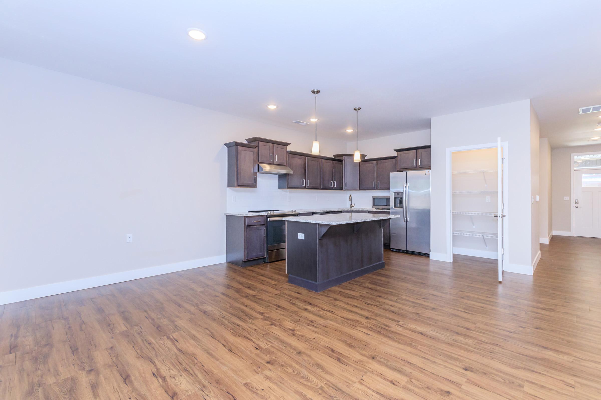 Modern open-concept kitchen and living area featuring dark wood cabinetry, a large island with a marble countertop, stainless steel appliances, and warm wooden flooring. Natural light fills the space from nearby windows, while a pantry is visible in the background.