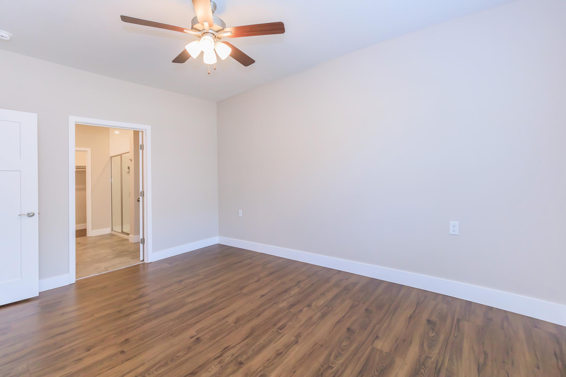 Interior of a room with a ceiling fan, light-colored walls, and a wooden floor. A doorway leads to a bathroom, visible in the background. The room is empty and well-lit, creating a spacious feel.