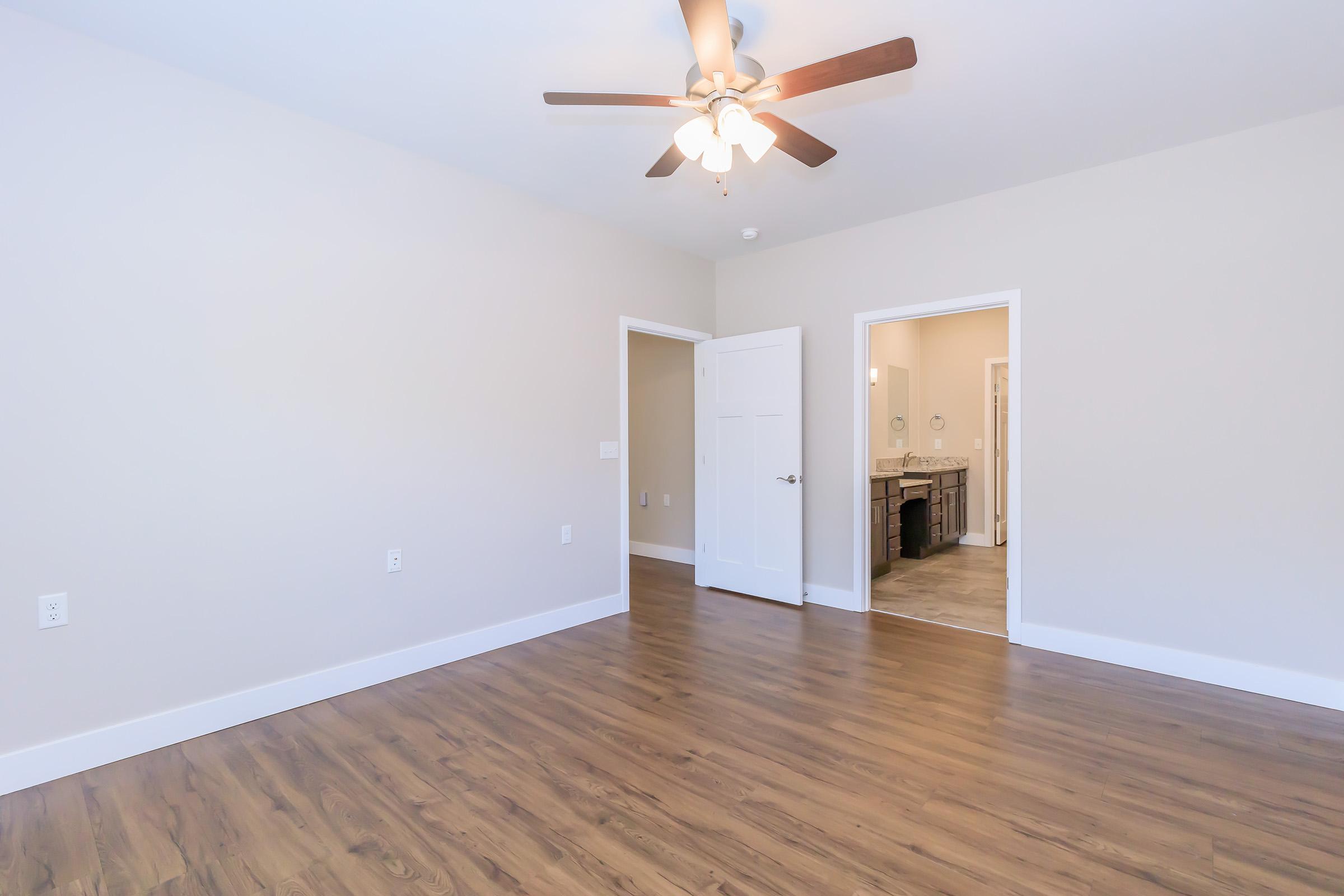 A spacious, empty room featuring light-colored walls and a ceiling fan. The floor is made of smooth wood, and there are two open doorways leading to adjacent areas. Natural light fills the space, creating a bright and inviting atmosphere. Ideal for various uses as a living area or office.