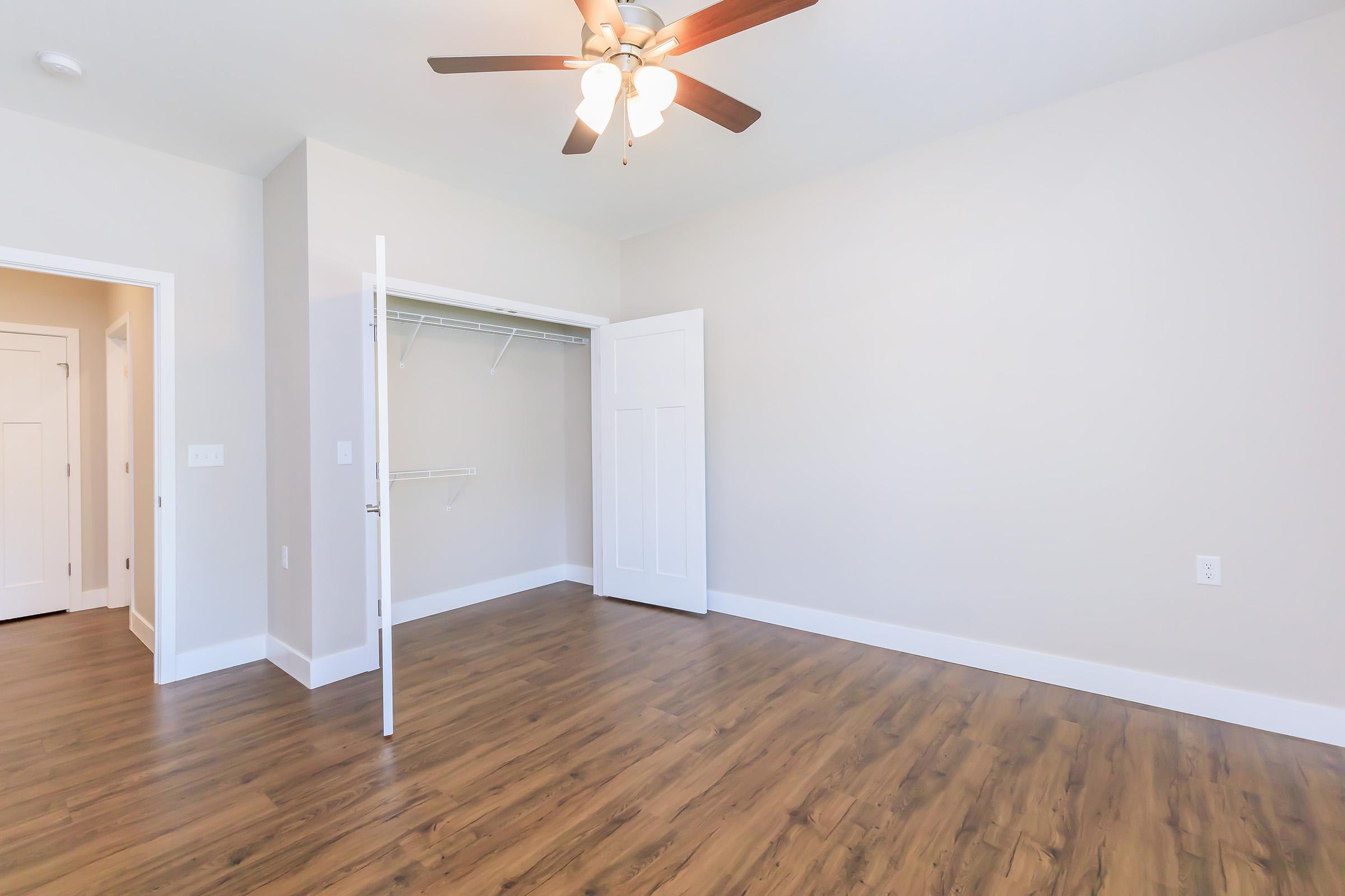 A bright, empty room with light-colored walls and a wooden floor. It features a ceiling fan with five blades, a closet with an open door, and a wide entryway. The room is well-lit and spacious, ideal for various uses such as a bedroom or home office.