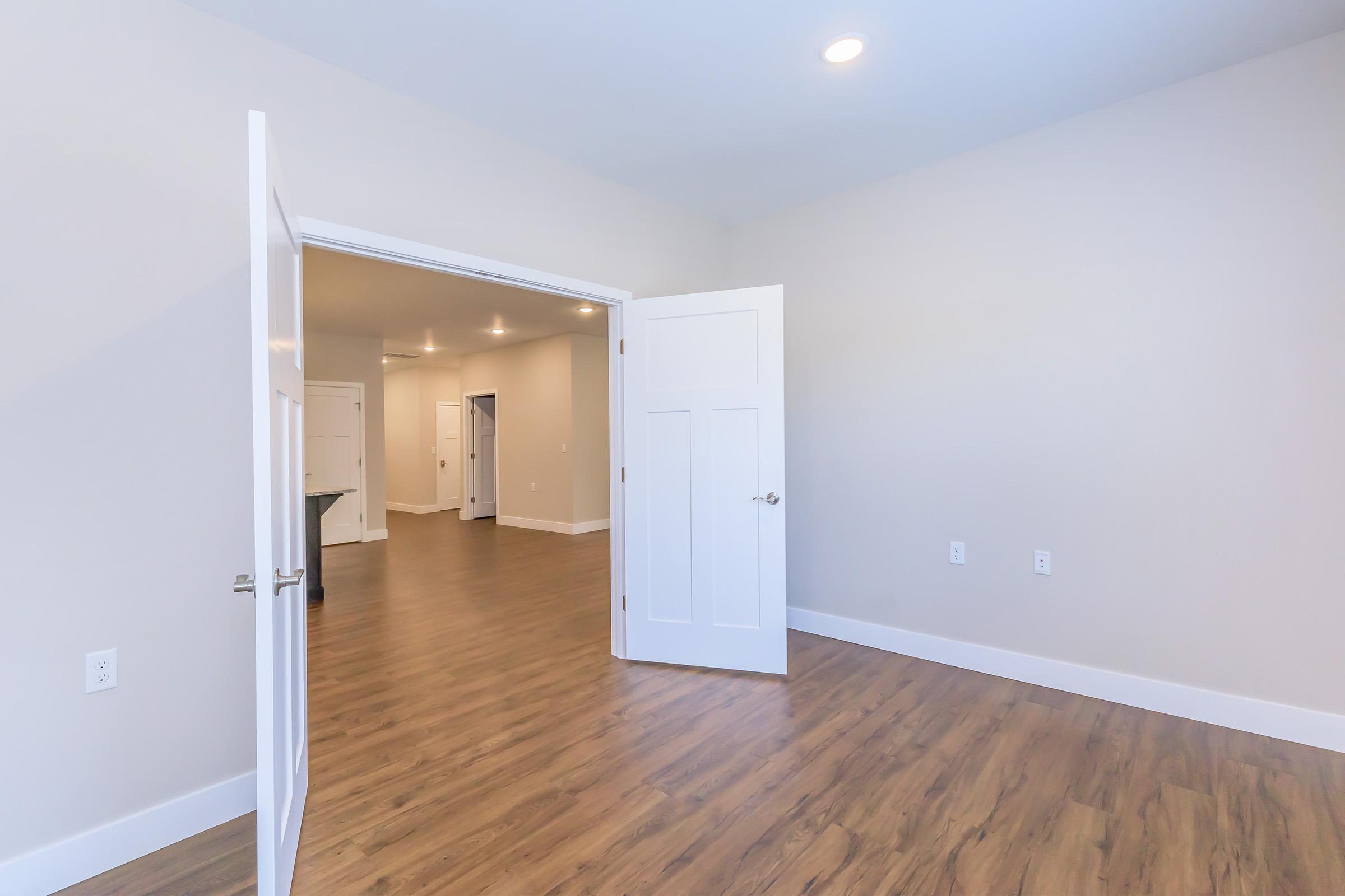 Interior view of a bright room with light-colored walls and a wooden floor. An open door leads to another room in the background, which has white walls and a modern design. The space is well-lit with recessed lighting, creating a clean and inviting atmosphere.