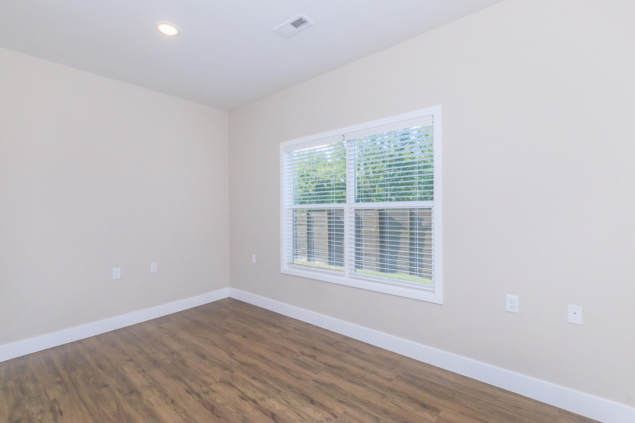 Empty room with light-colored walls, wooden flooring, and a window with white blinds allowing natural light to enter. The window offers a view of the outdoors, while the space is unfurnished and has minimal decor.