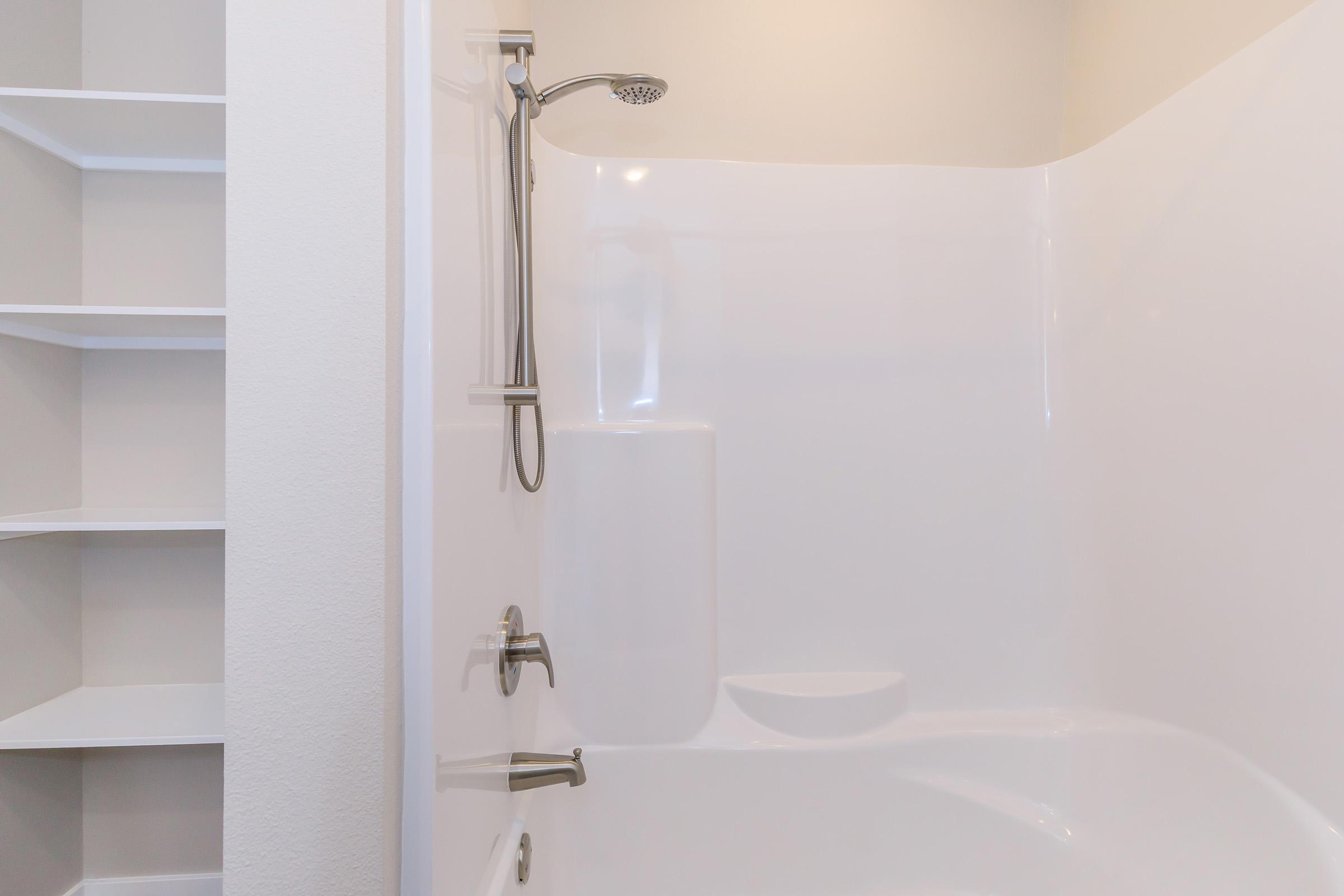 A clean and modern shower area featuring a wall-mounted showerhead and a fixed tub spout. The surrounding walls are smooth and white, with a low ledge for toiletries. To the left, a built-in shelf provides storage space. The overall design is minimalistic and bright, creating an inviting atmosphere.