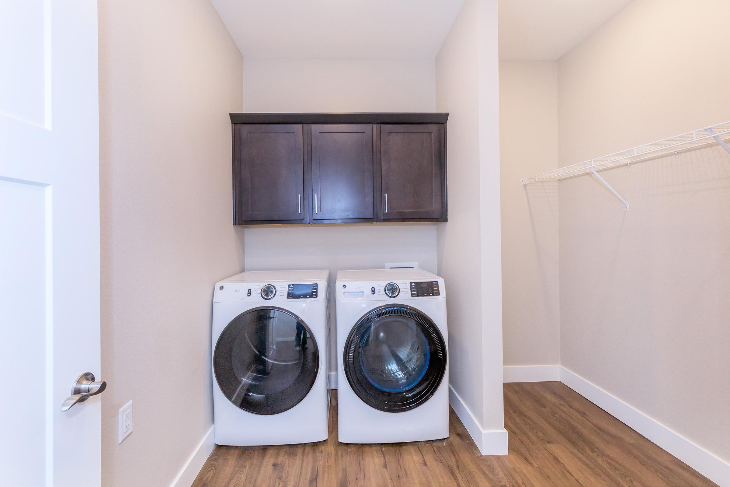 A modern laundry room featuring a stacked washer and dryer in white, placed next to dark wooden cabinets. The room has light-colored walls and a light wooden floor, with a wire shelving unit on one side for additional storage.