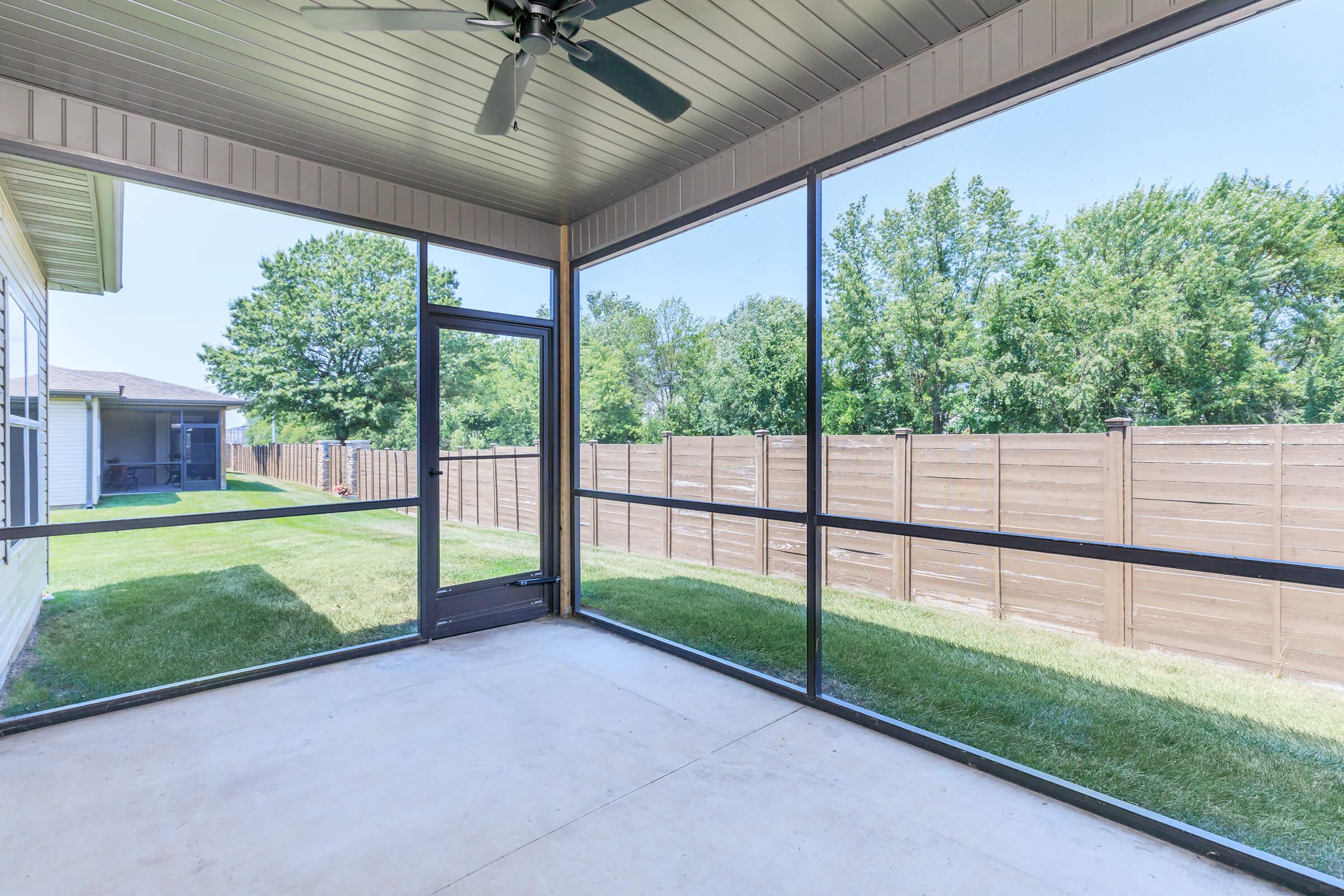 A spacious screened-in porch with a ceiling fan, featuring a concrete floor and expansive views of a green lawn and a wooden fence in the background. Sunlight filters through the screens, creating a bright and inviting atmosphere for relaxation.