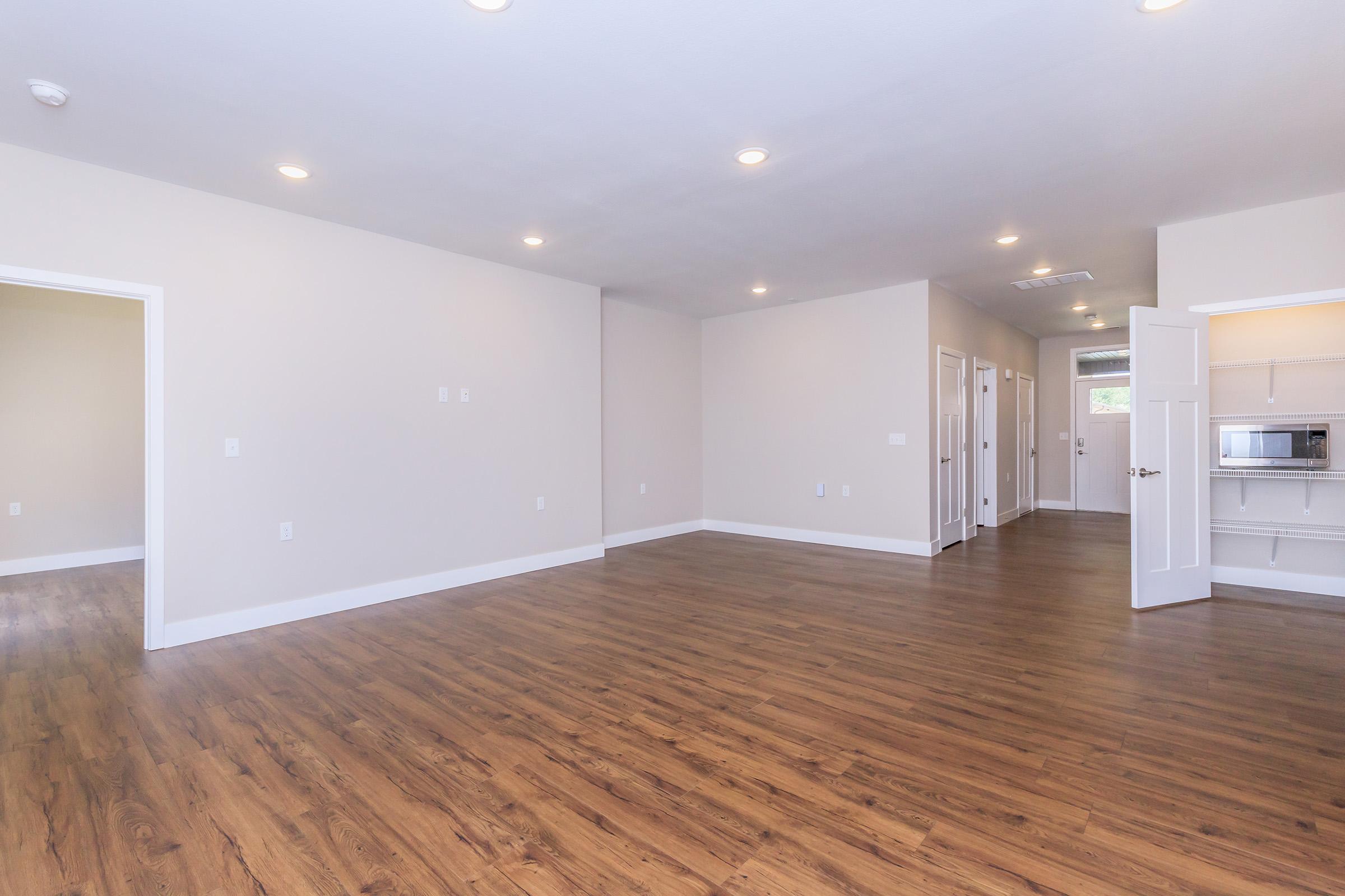 Spacious interior of an empty room with light-colored walls and warm wooden flooring. The room features multiple doorways leading to adjacent spaces, recessed lighting on the ceiling, and a large window allowing natural light. Ideal for different layouts or renovations.