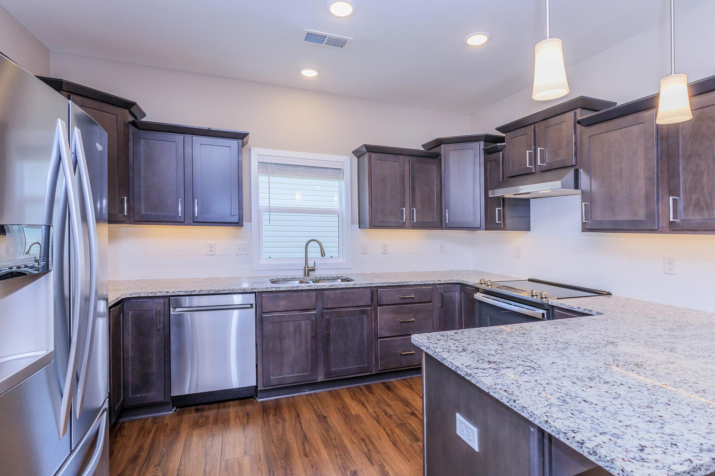 Modern kitchen featuring dark wood cabinetry, stainless steel appliances including a refrigerator and dishwasher, a granite countertop, and pendant lighting. A large window provides natural light, and the space has an open layout with a wooden floor.