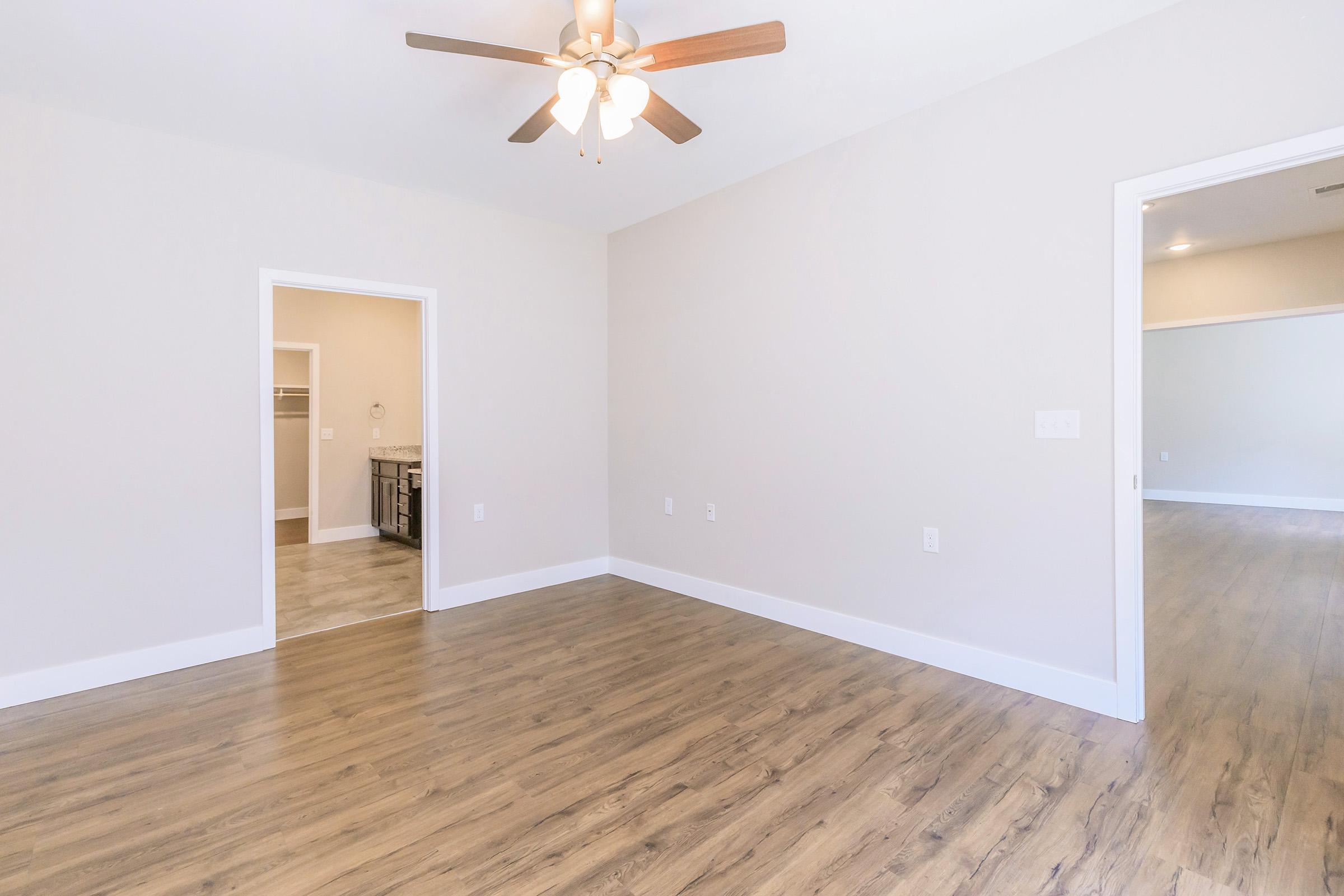 A spacious, empty room featuring a ceiling fan, light wood flooring, and neutral-colored walls. There are two doorways leading to other areas, with one doorway opening to a bathroom or closet space. The room is well-lit with natural light, creating an inviting atmosphere.