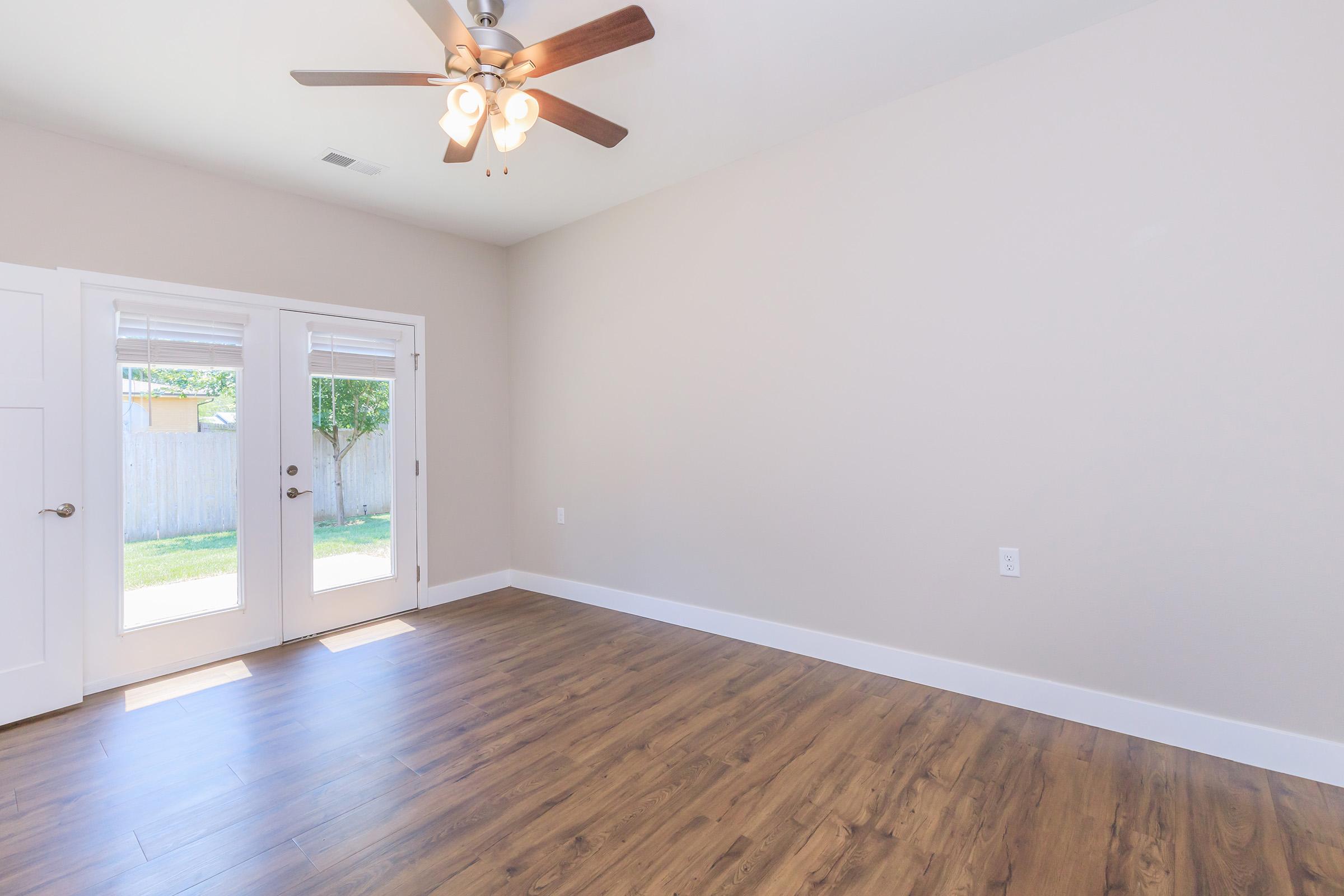 Spacious, unfurnished room featuring a ceiling fan, large windows with natural light, and neutral-colored walls. Double doors open to an outdoor space, while the floor is laid with wood-like laminate. Ideal for various uses such as a living area or office.