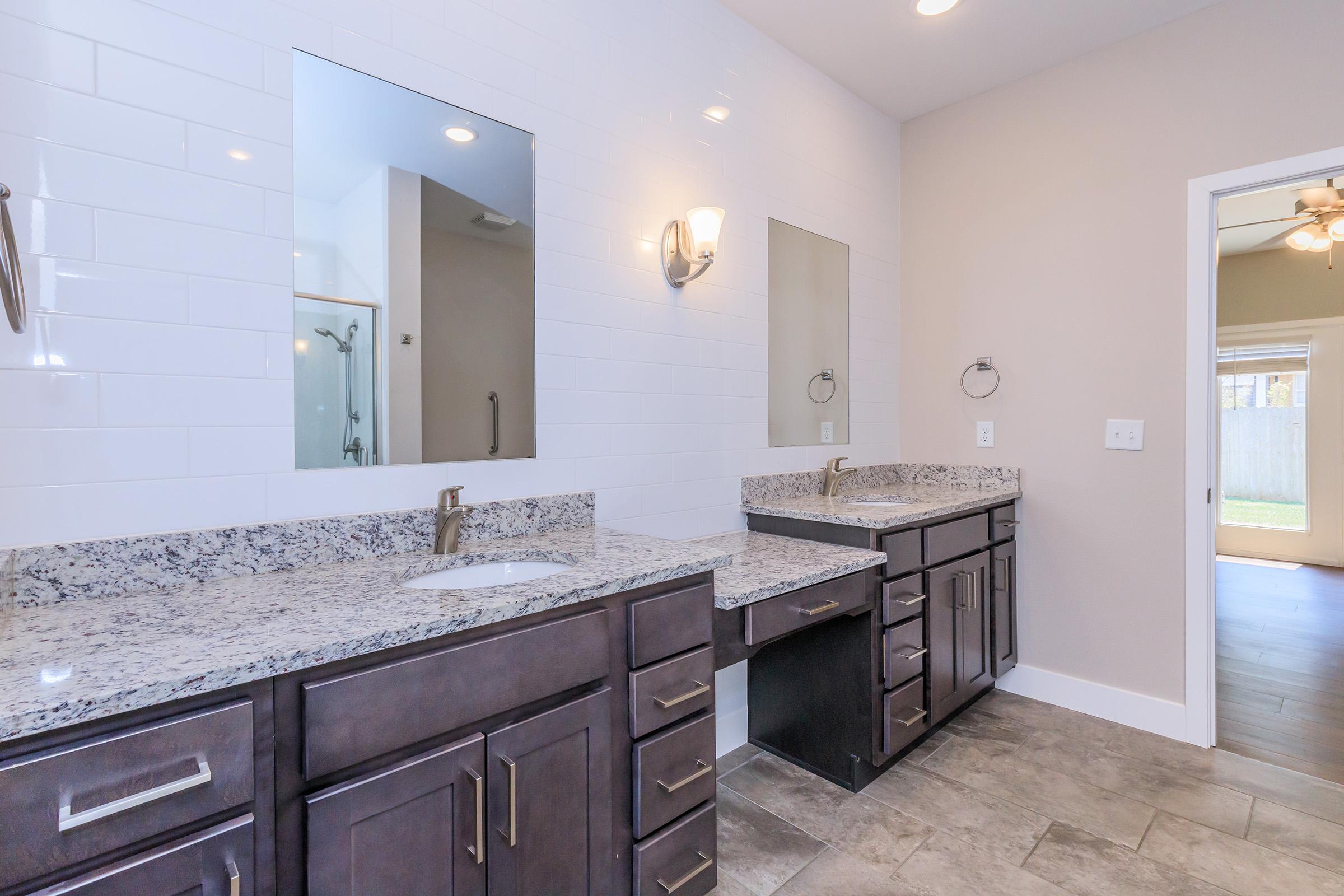 A modern bathroom featuring dual vanities with granite countertops, two mirrors, stylish wall sconces, and a light gray tiled floor. The spacious room has a shower area visible in the background, with a doorway leading to another room. Natural light fills the space, enhancing its contemporary design.