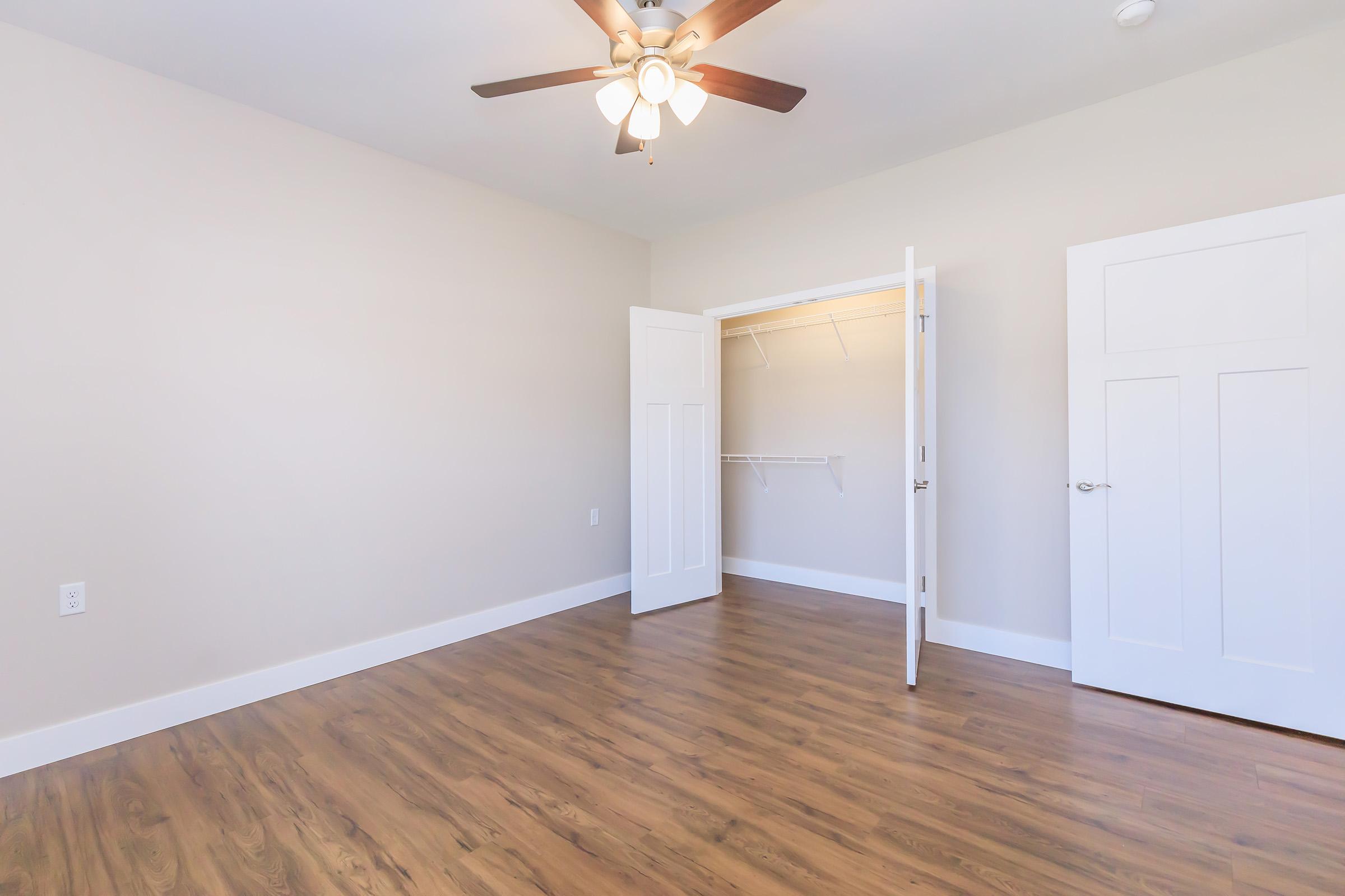 Empty room with light-colored walls and a ceiling fan. The floor is made of brown wood laminate. A closet is visible with white doors, slightly open, revealing shelves inside. The overall space is bright and uncluttered, providing a minimalist look.