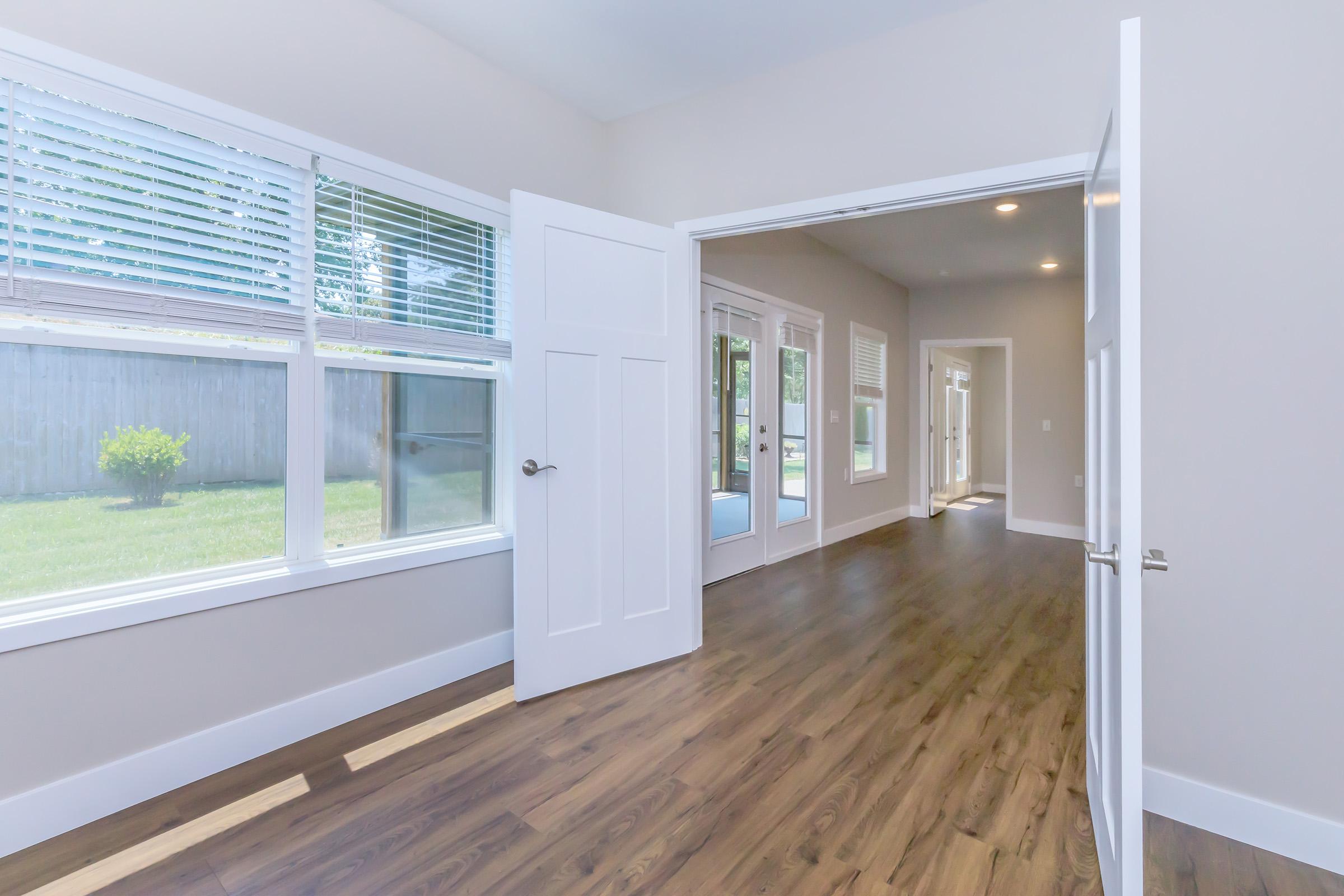 Bright and airy interior of a modern home featuring open doorways, large windows, and wood flooring. Natural light floods the space, creating a welcoming atmosphere. The room leads to another area with additional windows and green yard visible outside.