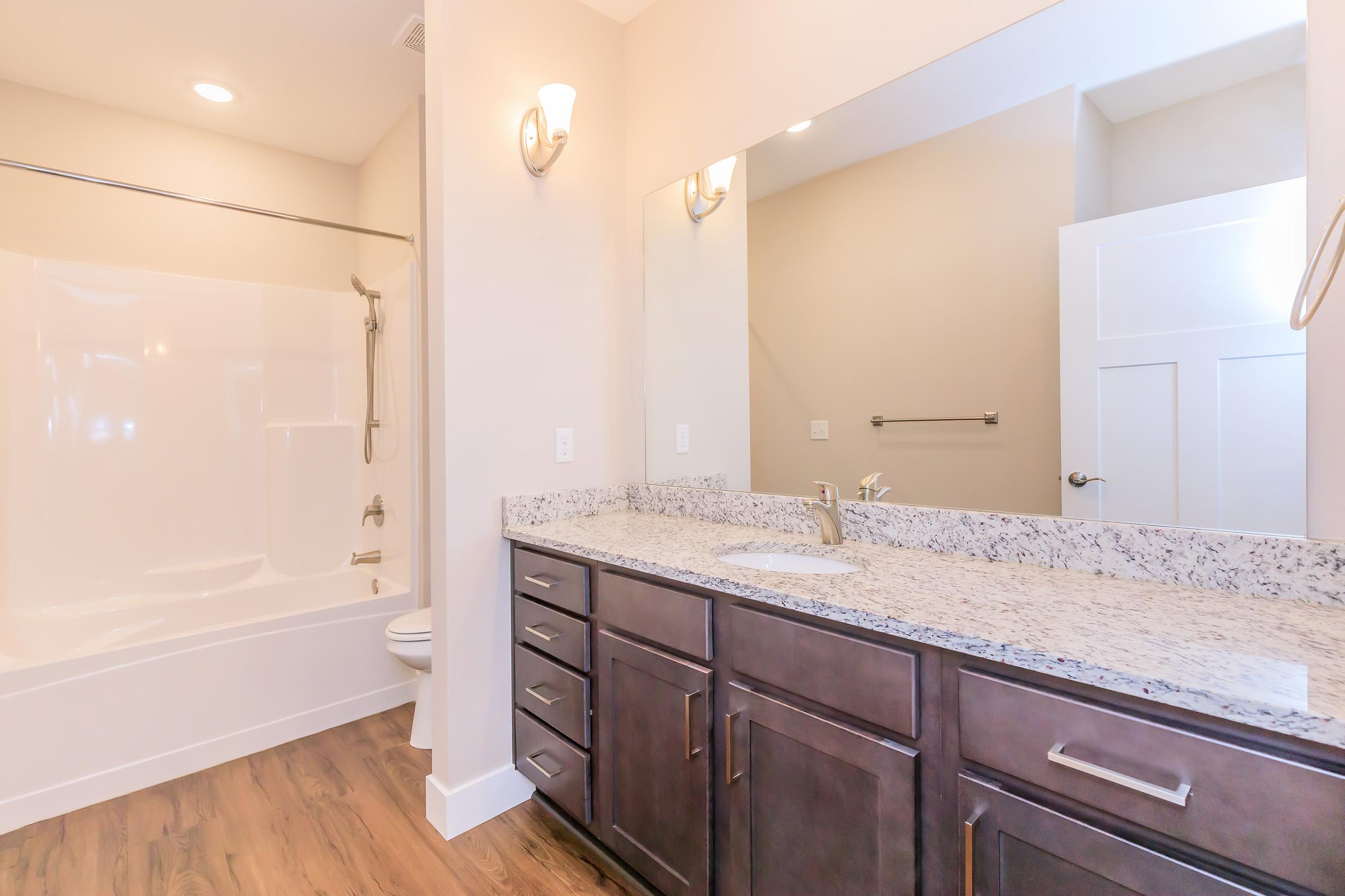 A modern bathroom featuring a light-colored granite countertop with a sink, dark wood cabinets, and a large mirror. There is a white bathtub with a showerhead, a toilet, and light-colored walls, creating a clean and spacious appearance. Natural light enters through a nearby window.