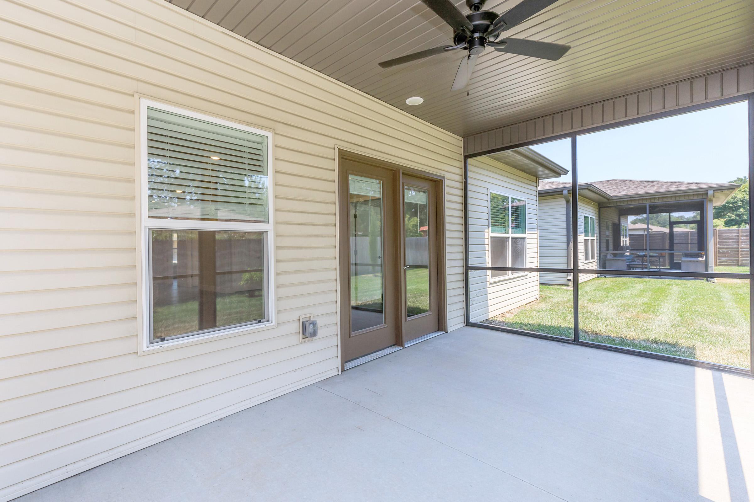 A screened-in porch featuring beige siding, a ceiling fan, and double doors leading to a backyard. The area is well-lit with large windows and offers a view of a grassy lawn and adjacent home, creating a cozy outdoor space.