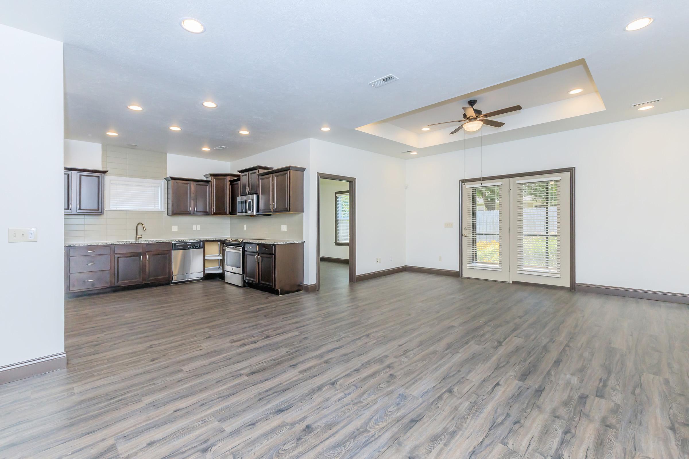 Spacious modern living area with a connected kitchen featuring dark wooden cabinets and stainless steel appliances. Large windows allow natural light to fill the room, which has a ceiling fan and is finished with light-colored walls and dark wood flooring. The layout creates an open and inviting atmosphere.